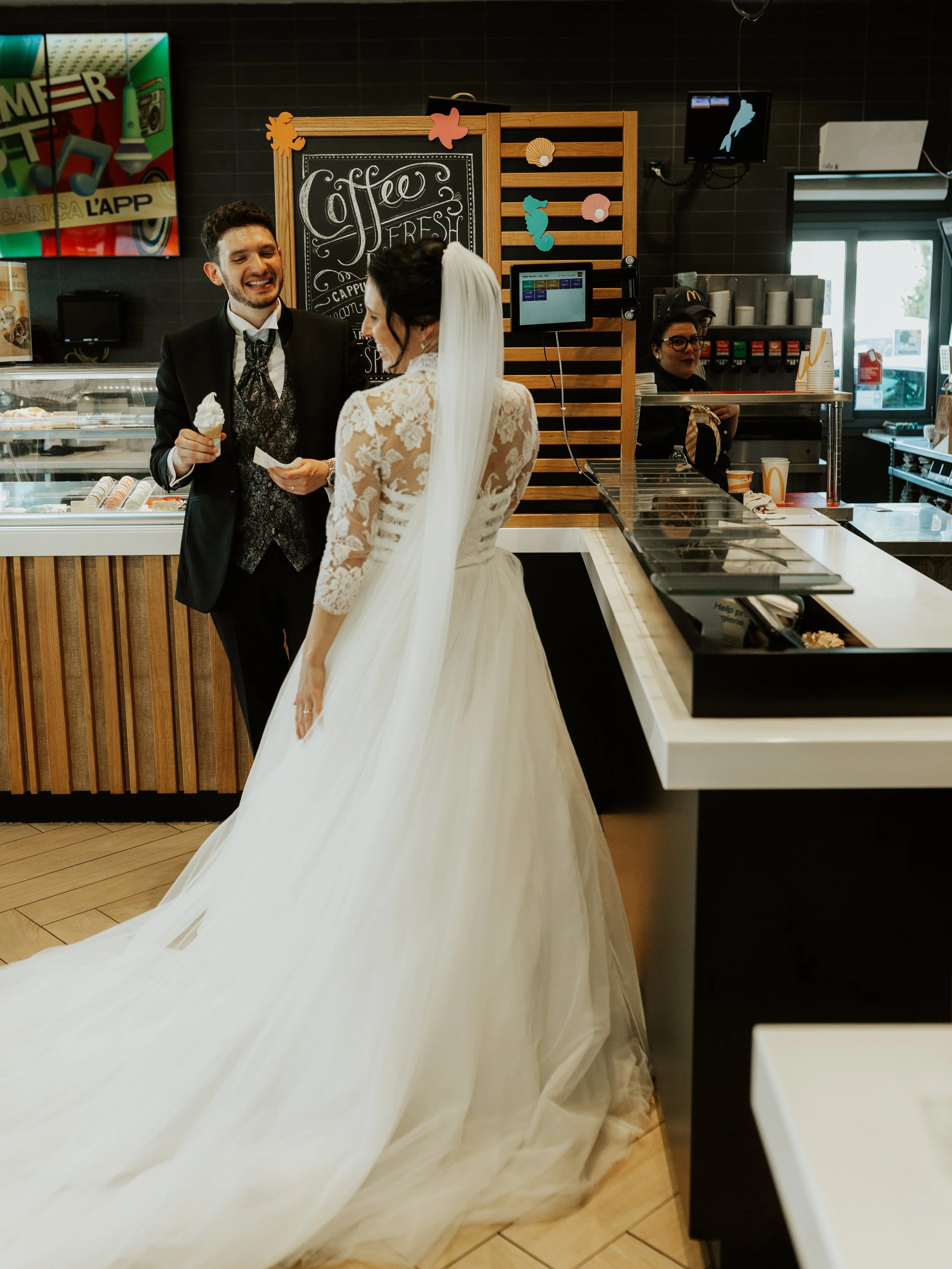 Una coppia di sposi in abiti da matrimonio sorride e parla all'interno di una gelateria.