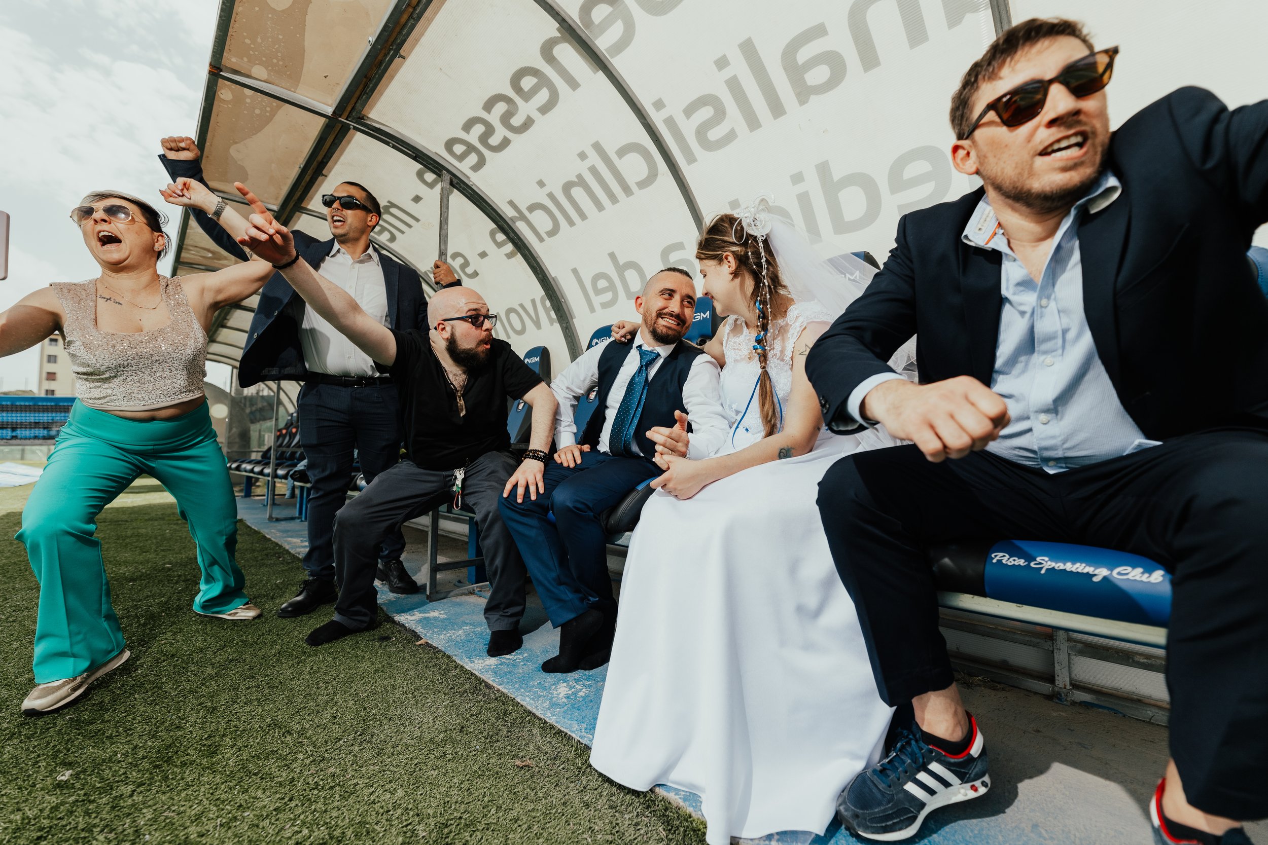 Gruppo di persone che si diverte sulla panchina di una tribuna allo stadio, con alcuni che sembrano discutere animatamente e altri che sorridono e si abbracciano.