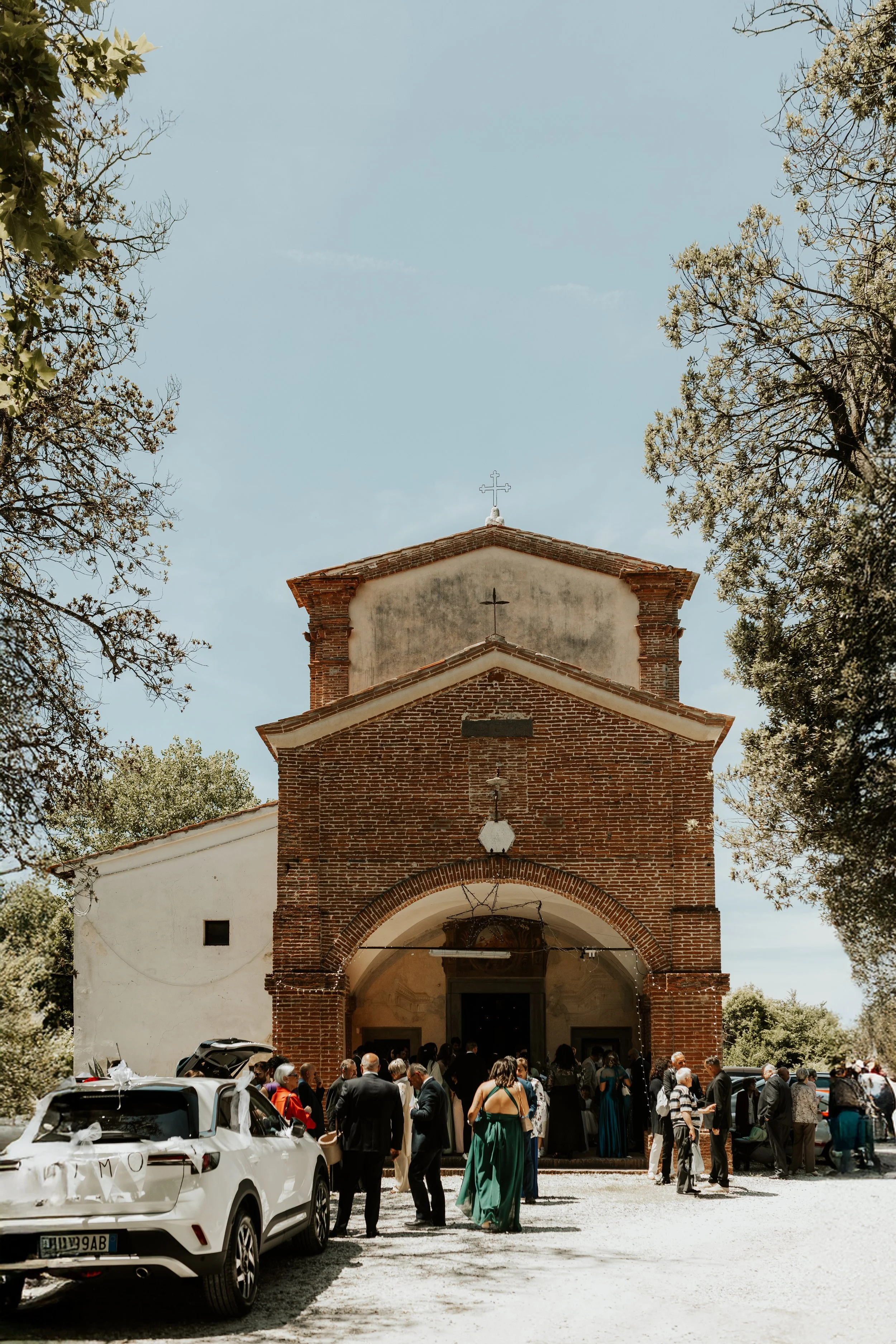 Chiesa in mattoni rossi con persone fuori, alcune in abiti eleganti, e auto parcheggiate davanti in un cortile con alberi intorno