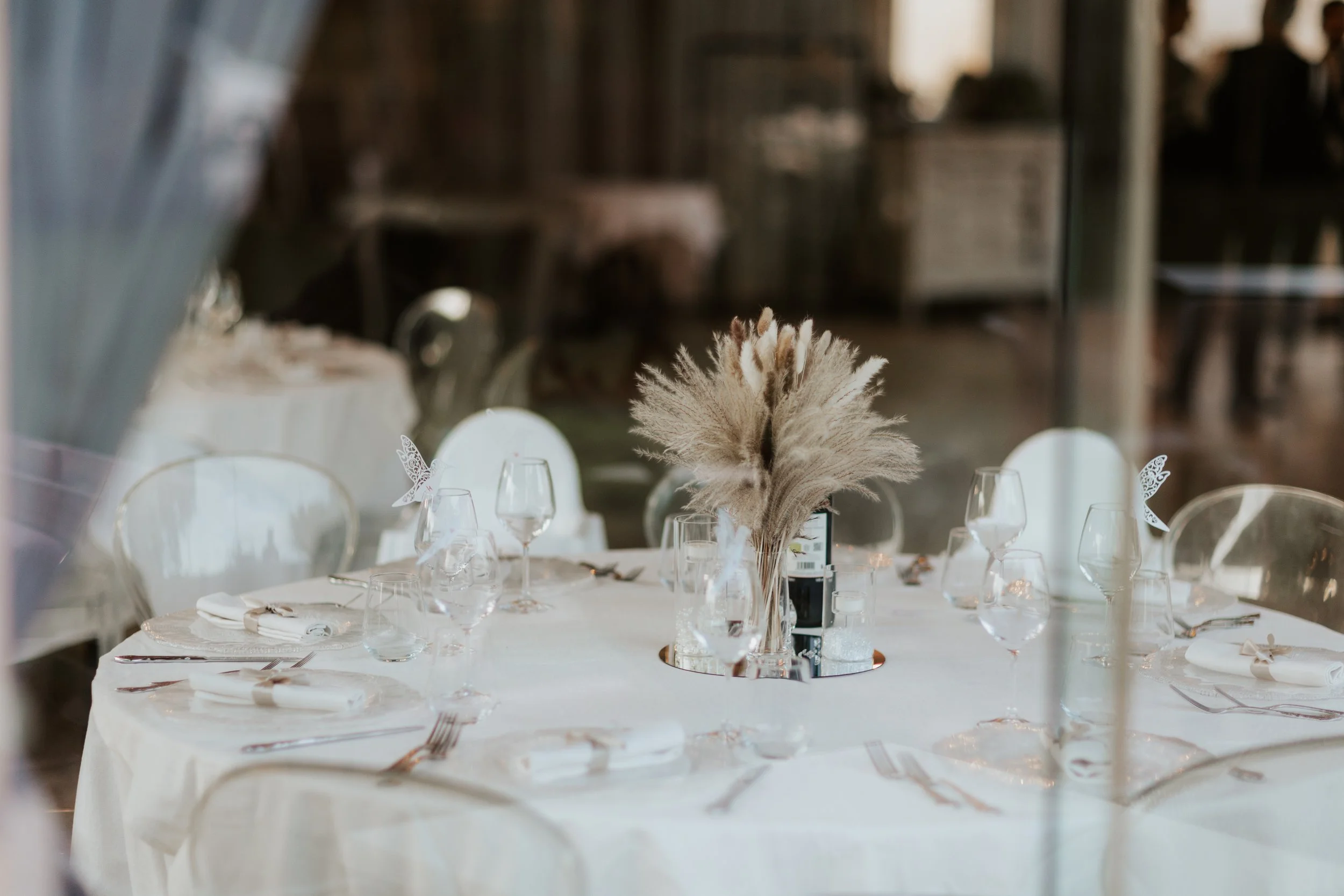 Tavolo apparecchiato per un evento con centrotavola di pampas e calici di vino, vista attraverso una finestra
