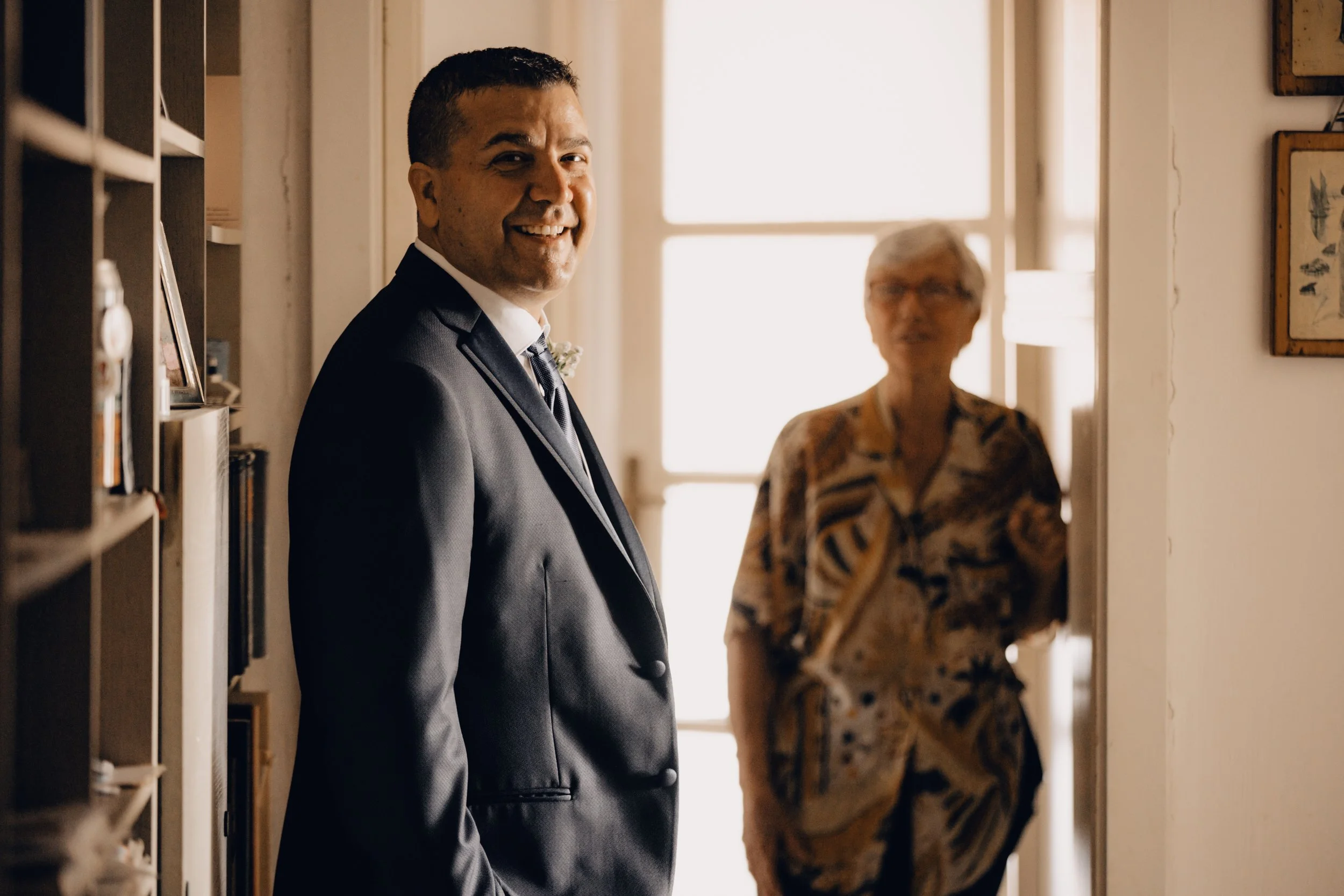 Uomo sorridente in abito elegante in un ambiente interno con una donna in sfondo.