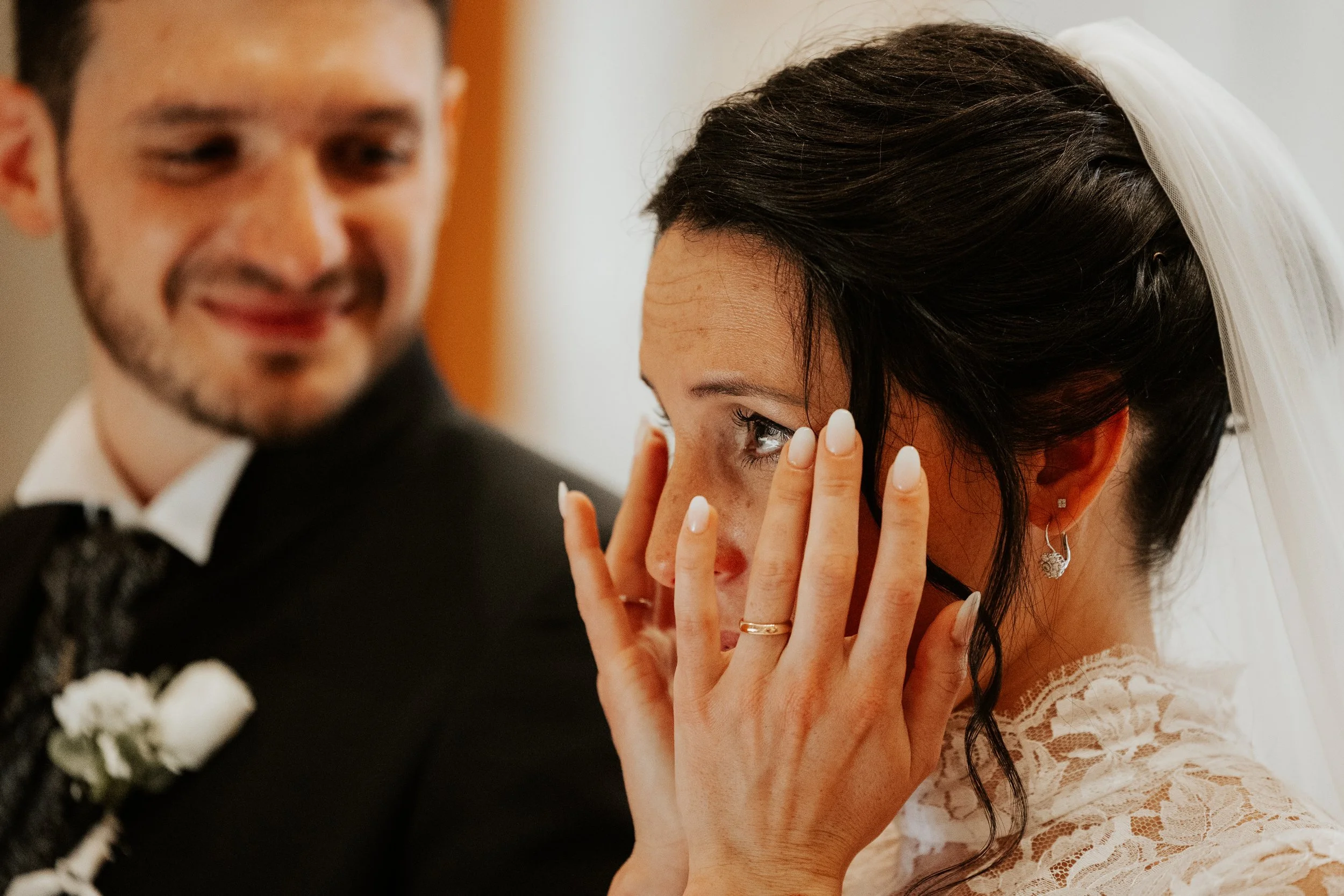 Una donna con capelli scuri e trucco leggero, coperta da un velo di nozze, è commossa e si sta asciugando le lacrime. Accanto a lei, un uomo sorridente, vestito elegantemente con giacca nera, camicia bianca e cravatta, è di fronte a lei durante un ma