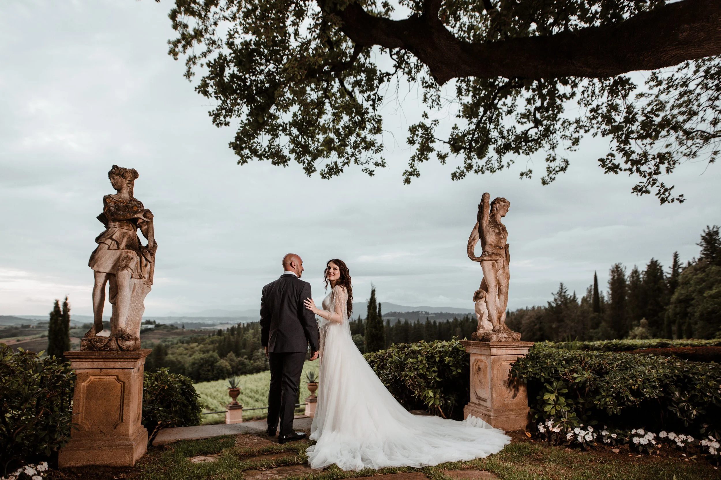 Coppia di sposi in abiti eleganti in un giardino con statue antiche, sotto un grande albero, con un paesaggio di colline e cielo nuvoloso sullo sfondo.