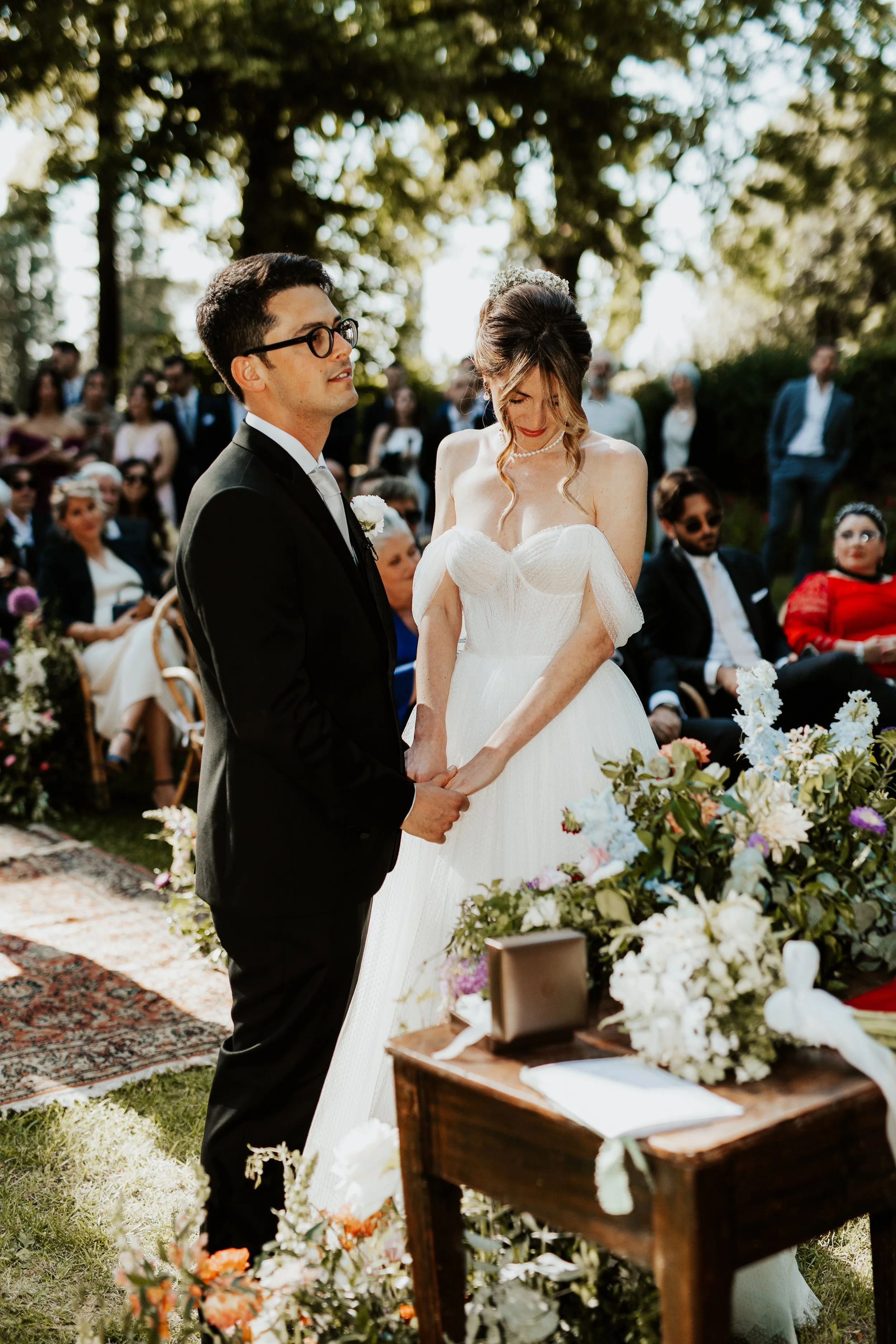 In matrimonio in un giardino, con lo sposo in abito nero e la sposa in abito bianco con scollo a cuore, mentre si tengono per mano durante la cerimonia. Villa dal Pozzo