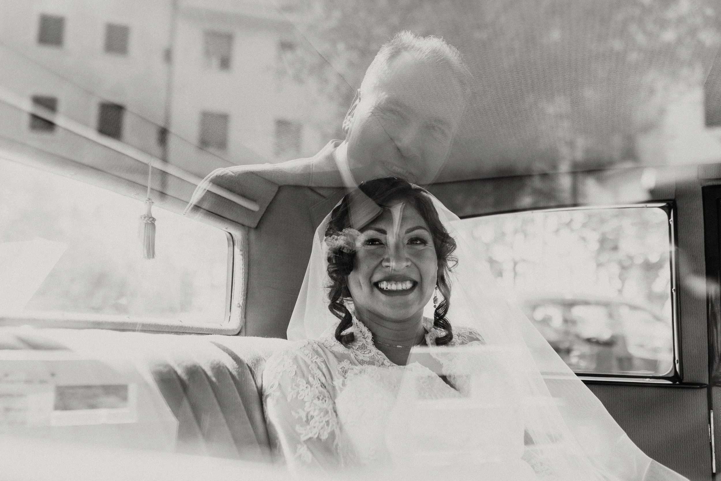 Donna sorridente in abito da sposa, seduta in un veicolo, visibile anche l'uomo in alto, probabilmente il suo fotografo o un ospite, con riflesso nel vetro dell'auto