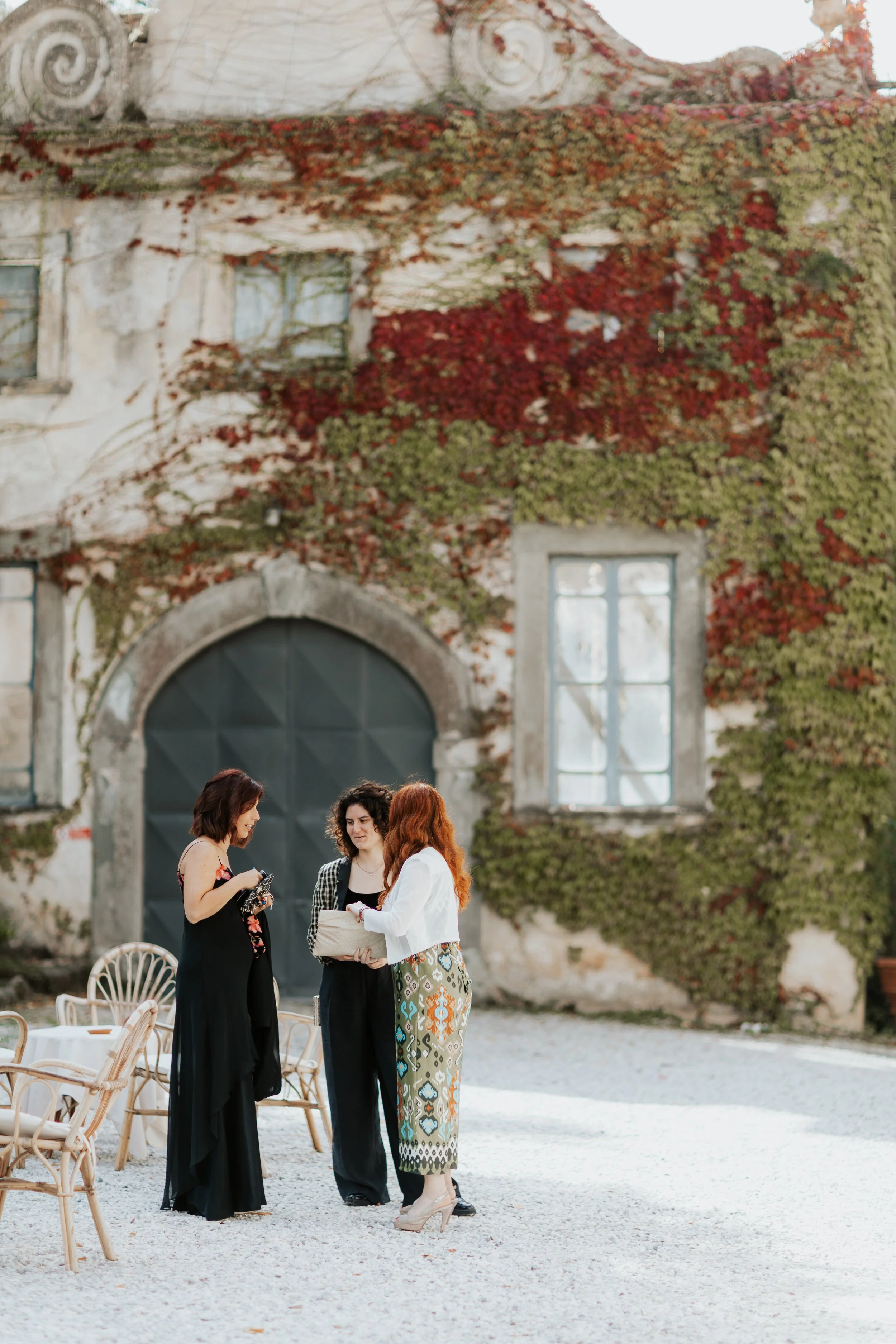 Tre donne che si incontrano e parlano all'aperto davanti a un edificio antico coperto di edera rossa e verde, con sedie di vimini sparse.