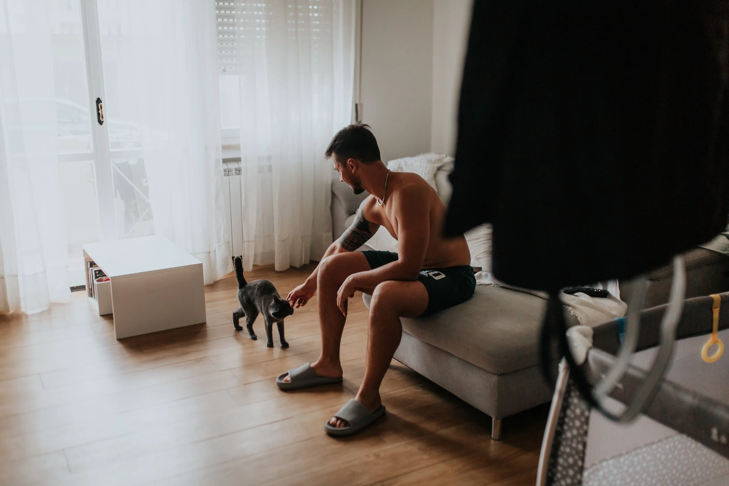 Un uomo senza maglietta e con pantaloncini scuri seduto su un divano in una stanza, dà da mangiare a un gatto nero che si avvicina a lui. La stanza ha grandi finestre con tende bianche e un pavimento di legno.