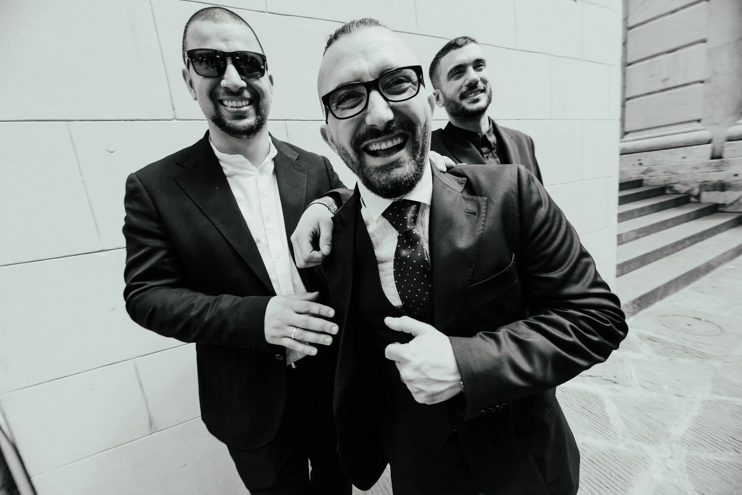Tre uomini sorridenti in abiti eleganti, uno con occhiali neri, uno con occhiali con cornice spessa e un con barba, che sorridono e si scambiano un abbraccio all'aperto.