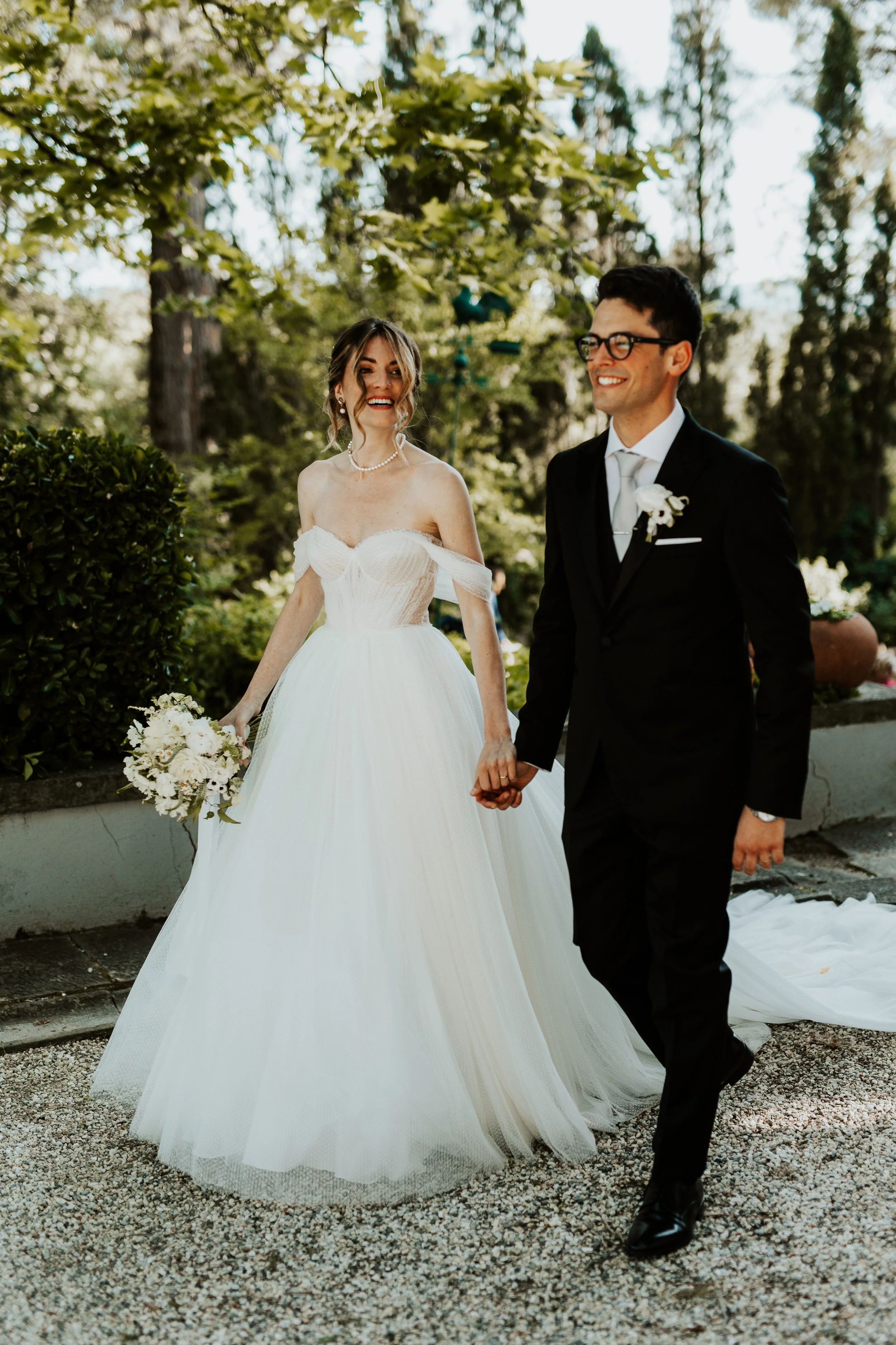 Una coppia di sposi si tiene per mano durante il matrimonio, con la sposa in abito bianco e sposa e lo sposo in completo nero, all'aperto tra gli alberi