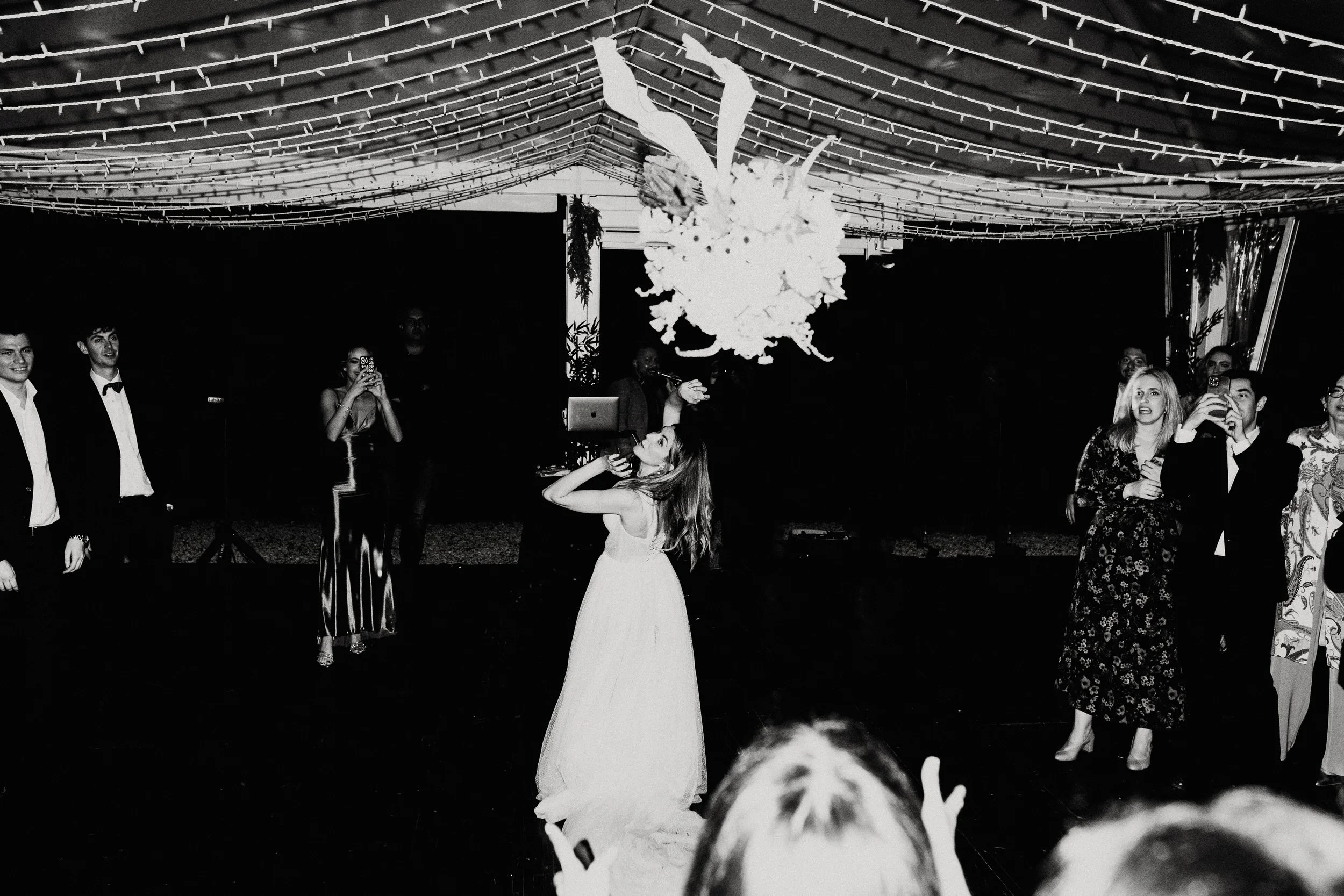 Una coppia di sposi balla al centro di una sala decorata con luci e un grande bouquet sospeso, mentre gli ospiti si guardarono e scattano foto durante il matrimonio.