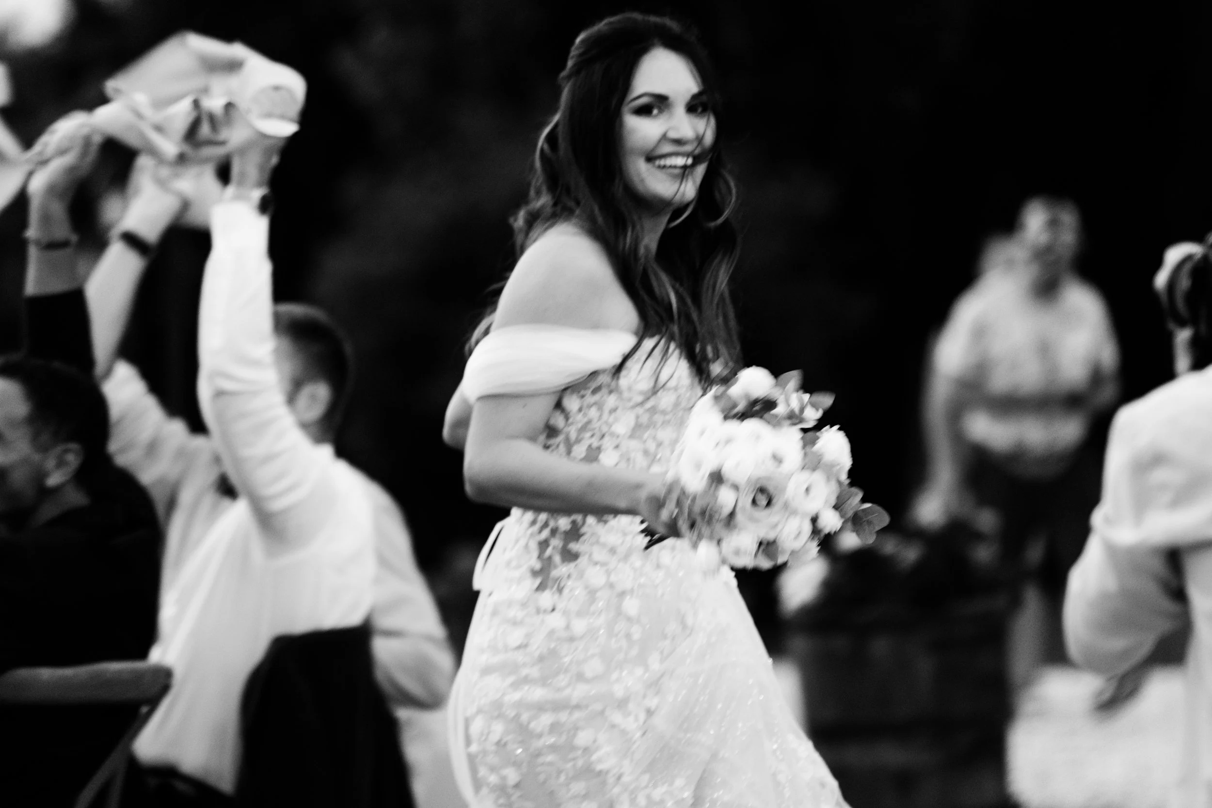 Una donna felice in abito da sposa con un bouquet di fiori, circondata da persone in un ambiente sociale, probabilmente a un matrimonio.