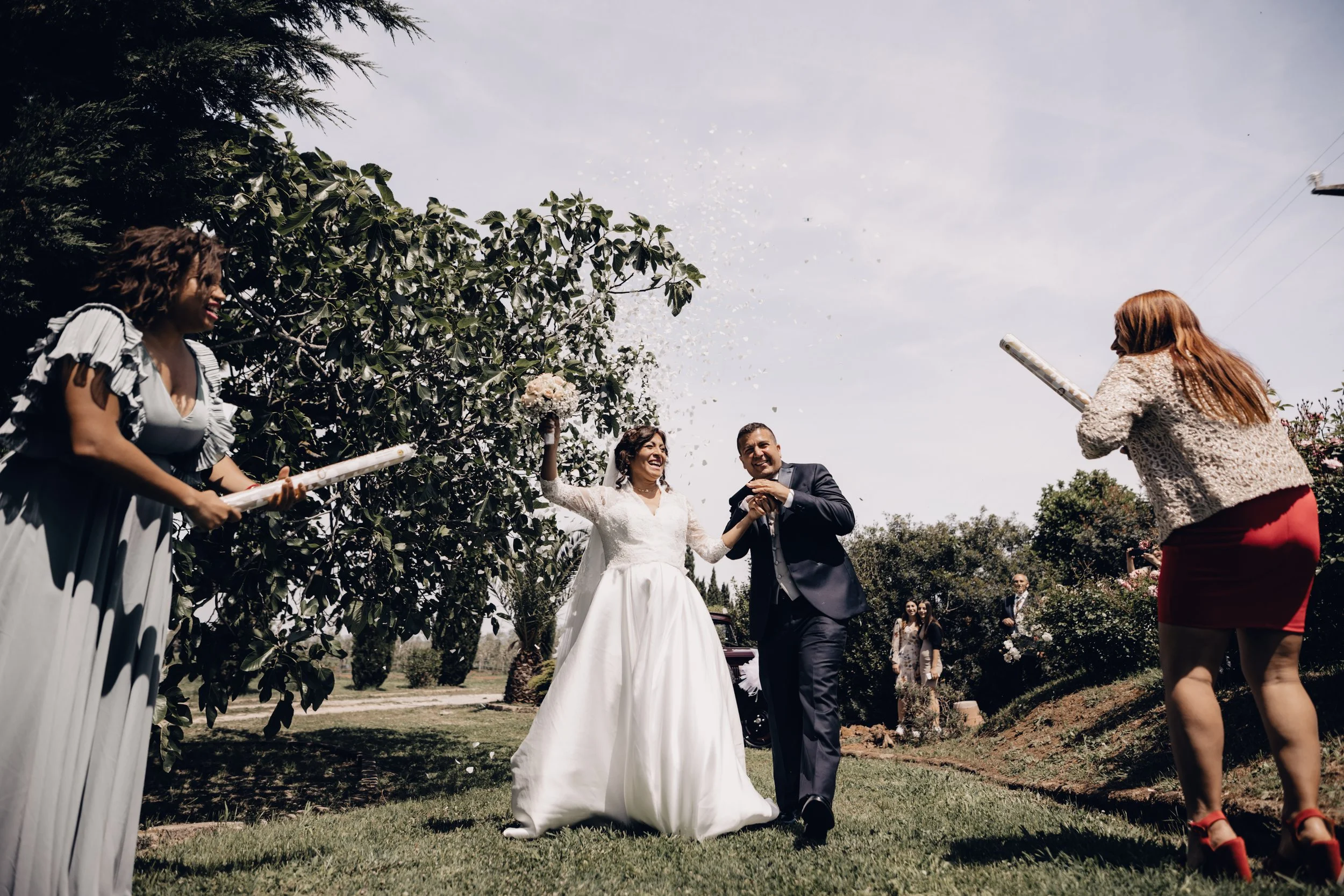 Una coppia di sposi festeggia con amici in un giardino durante un matrimonio, con momenti di allegria e confetti in aria.