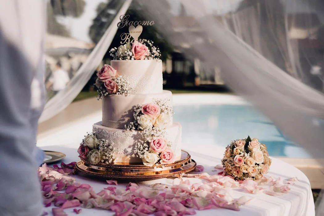 Torta nuziale a tre livelli decorata con rose rosa e bianche, fiori di baby's breath, su un tavolo con petali di rosa, vicino a un bouquet di rose.