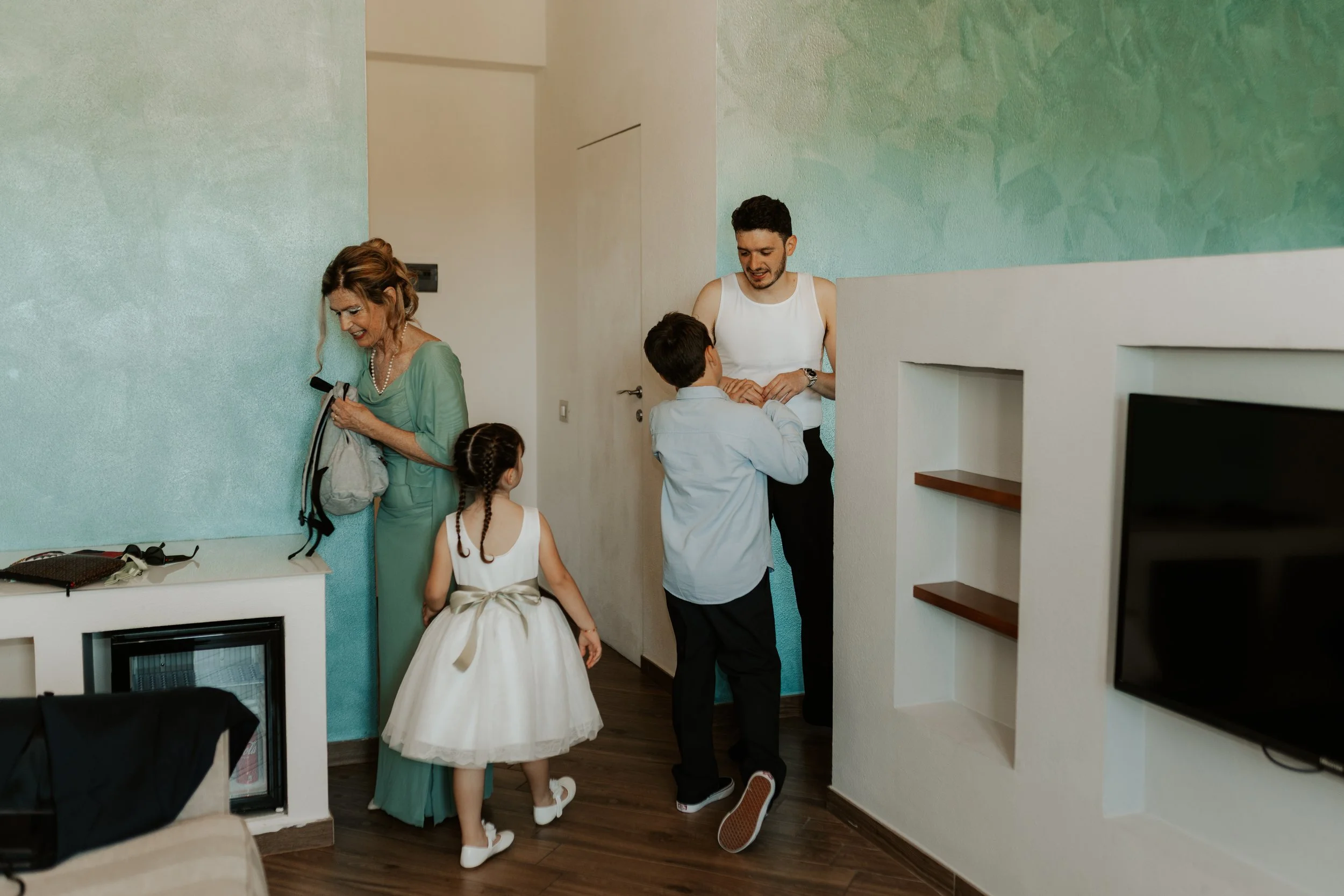 Famiglia in un soggiorno; donna anziana con borsa, giovane donna, due bambini, uno con vestito bianco e fiocco e l'altro con camicia bianca e pantaloni neri, stanno sorridendo e si salutano.