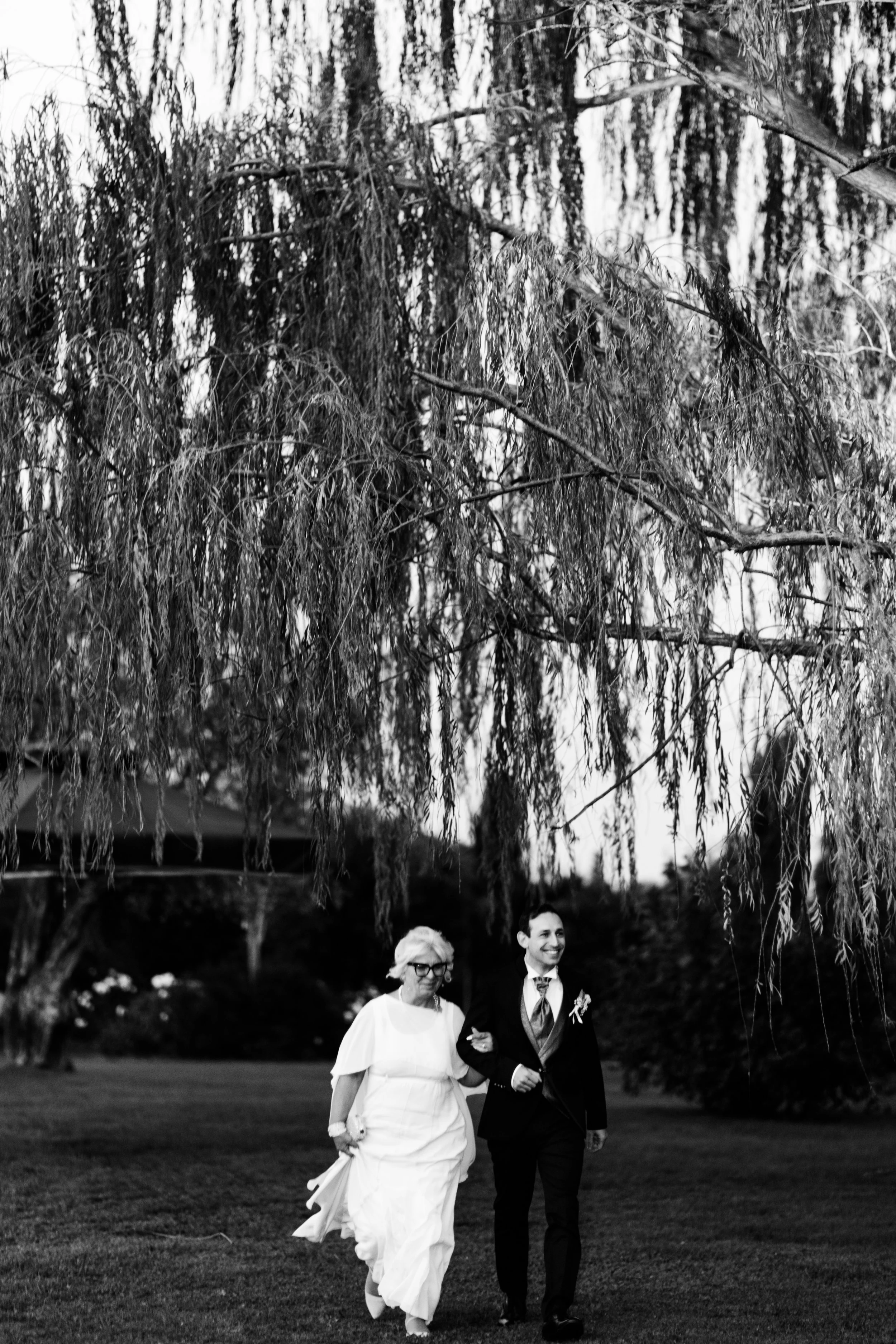 Foto in bianco e nero di due persone vestite da sposi che camminano sotto un albero ricadente. La donna indossa un abito bianco e occhiali, mentre l'uomo indossa un abito da uomo e una cravatta. Sono felici e si tengono per mano.