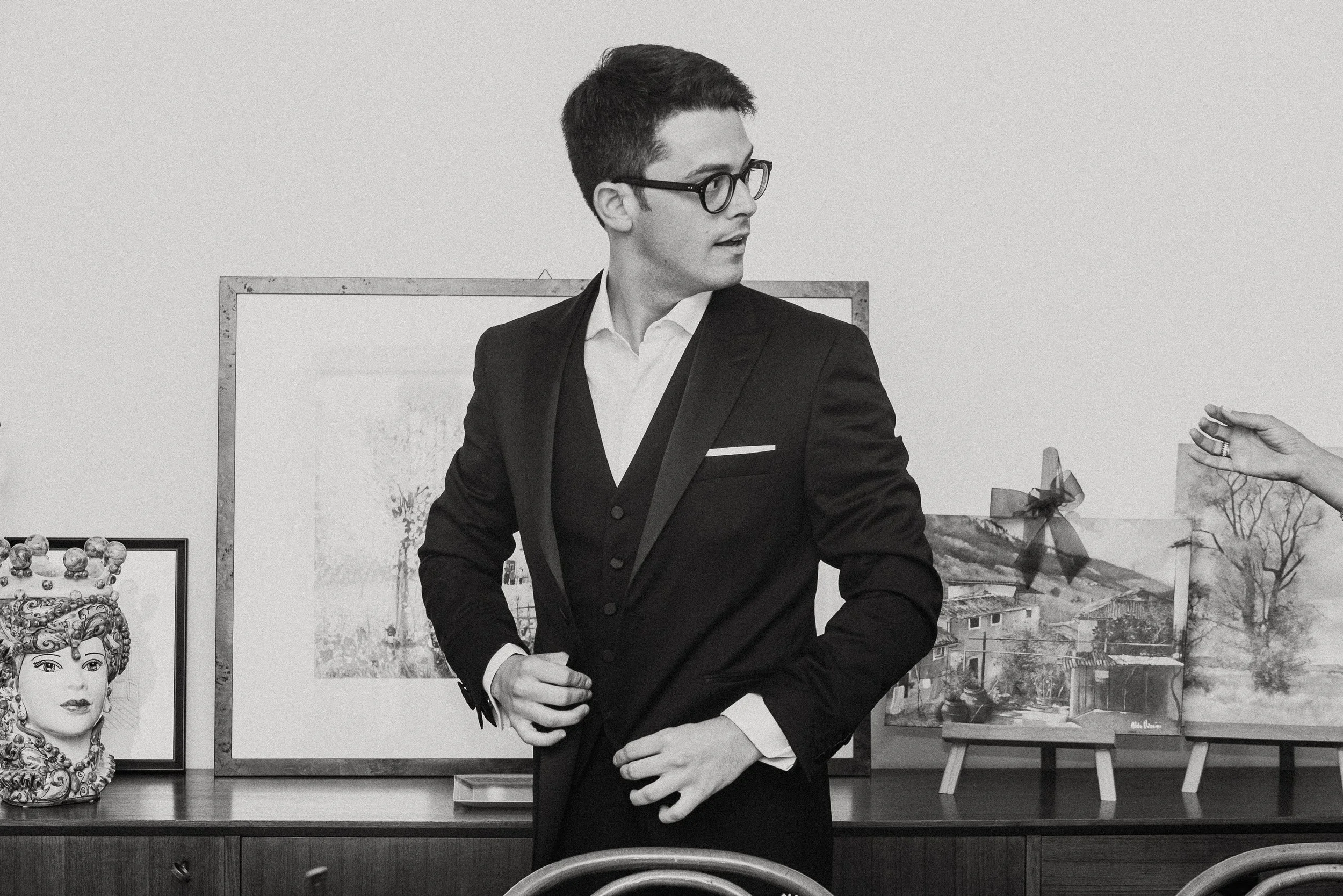 Un giovane uomo vestito con abito da sera nero, camicia bianca e occhiali da vista, in un ambiente con quadri e opere d'arte alle pareti