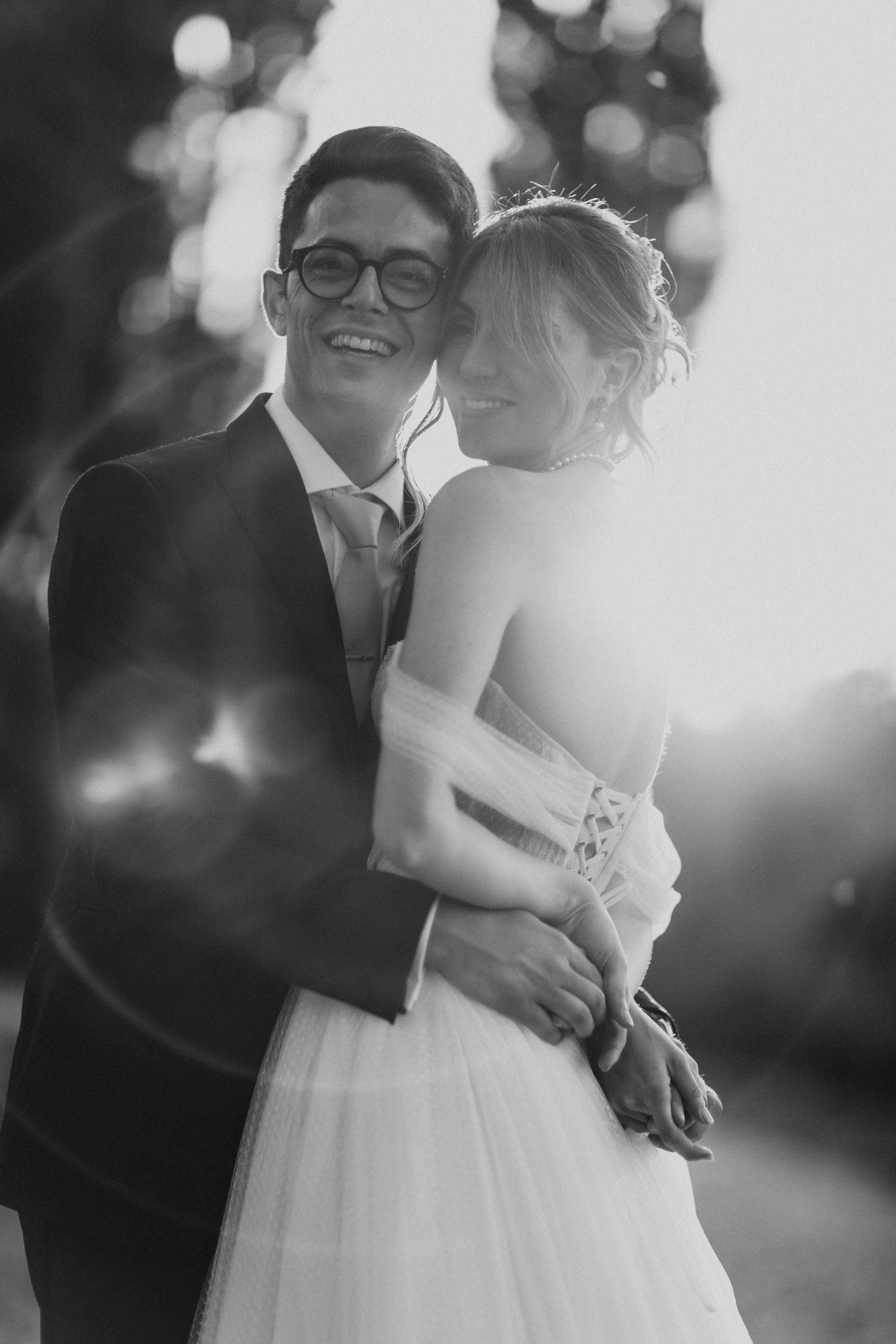 Una coppia di sposi sorride, vestita con abiti da matrimonio, in un soggetto fotografico in bianco e nero, con una forte luce di sfondo.