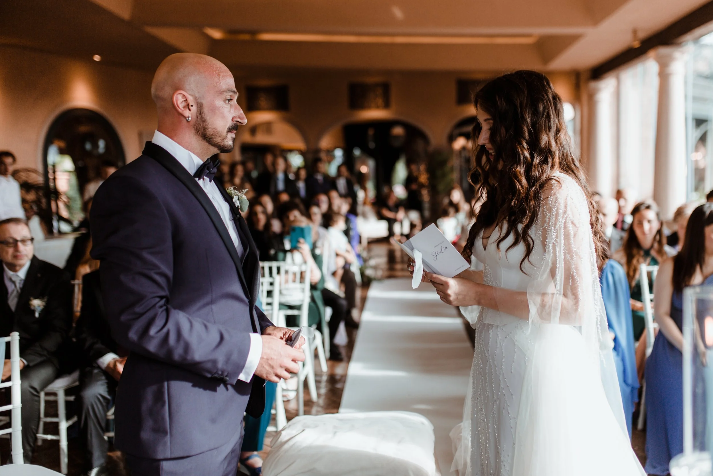 Sposo e sposa si lasciano leggere le promesse di matrimonio durante la cerimonia in una sala illuminata dalla luce naturale