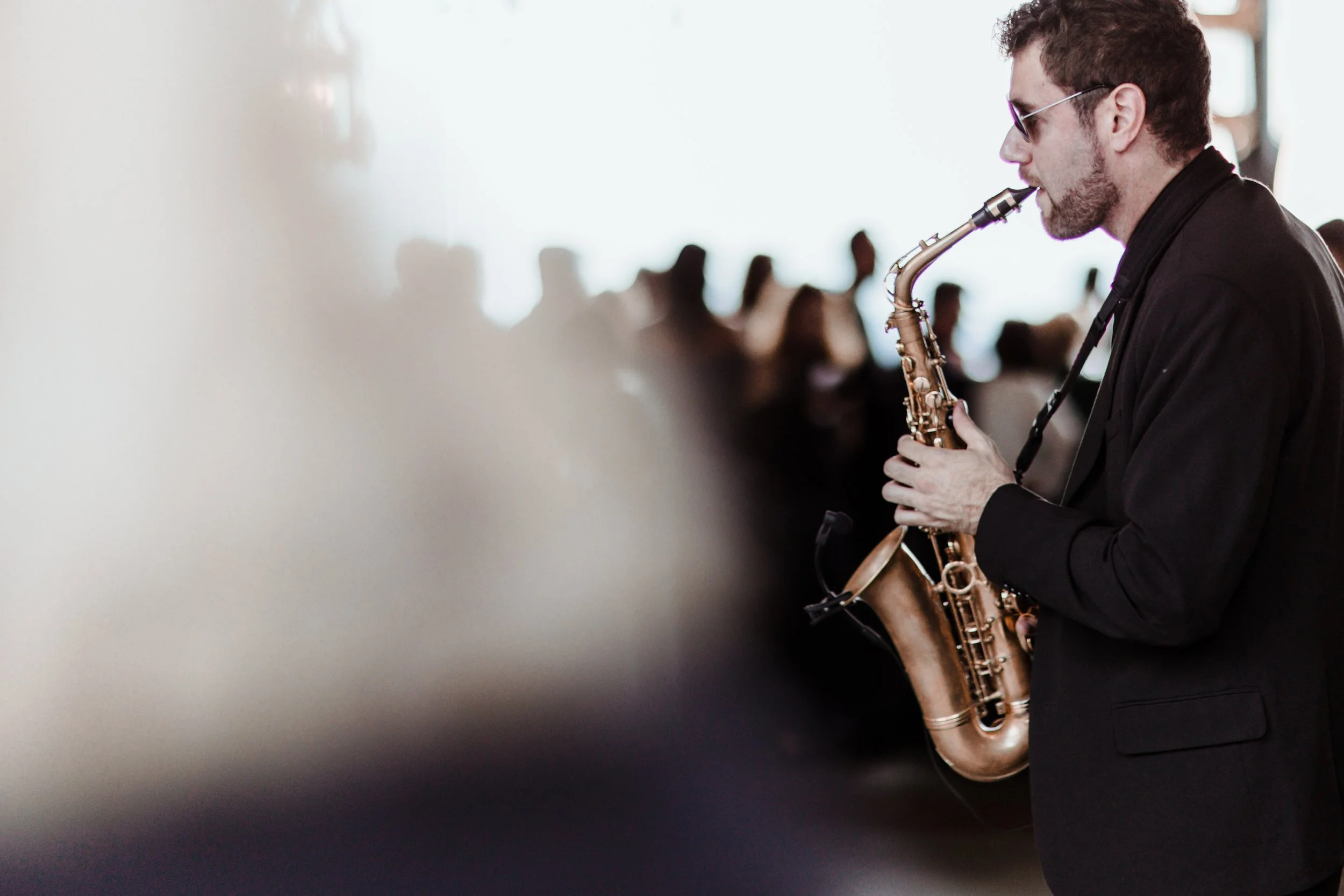 Uomo con occhiali da sole suona un sassofono in un ambiente con molte persone sfocate sullo sfondo.