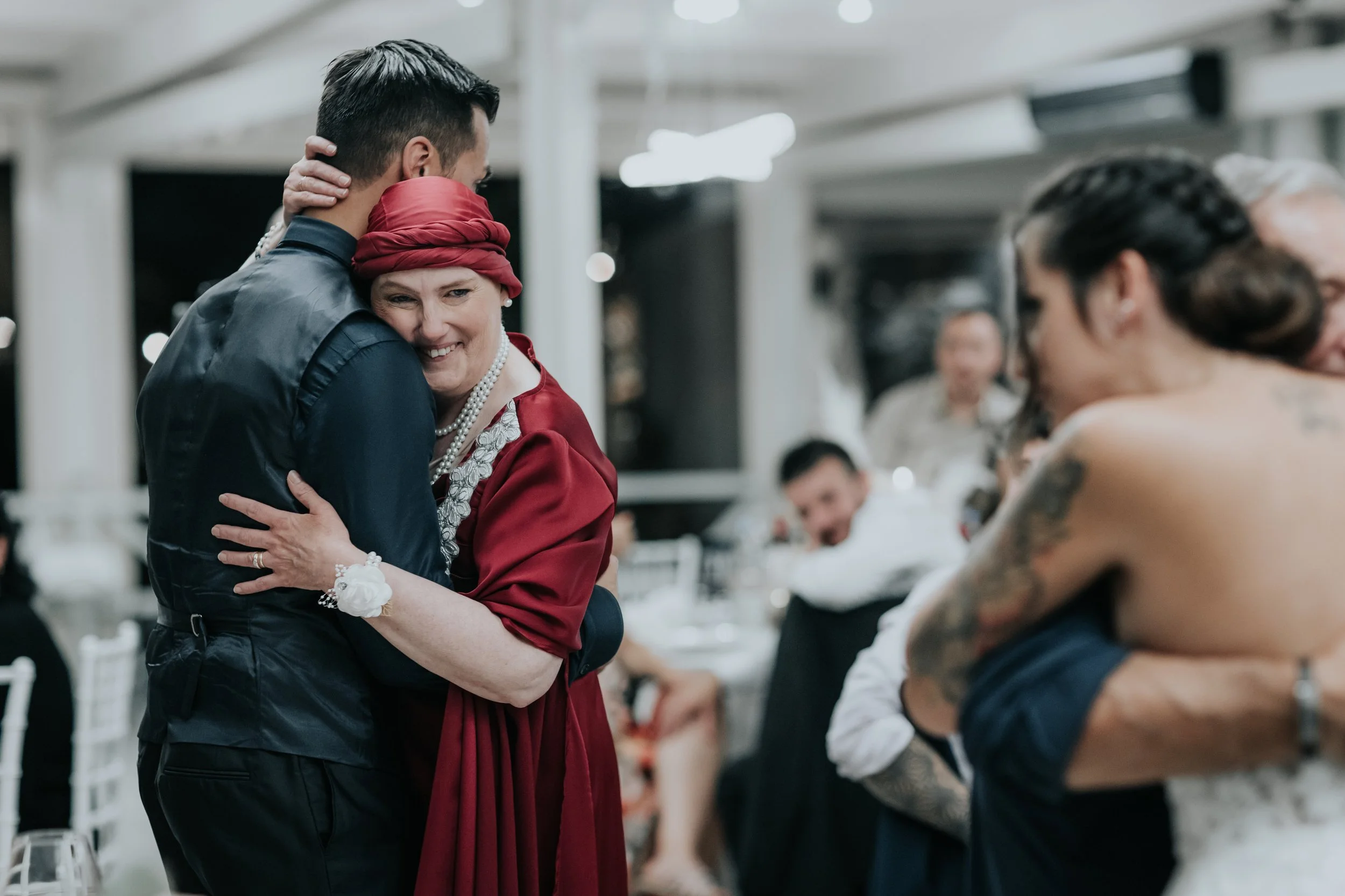 Persone che si abbracciano in un ricevimento, con uno sfondo di tavoli e altri ospiti