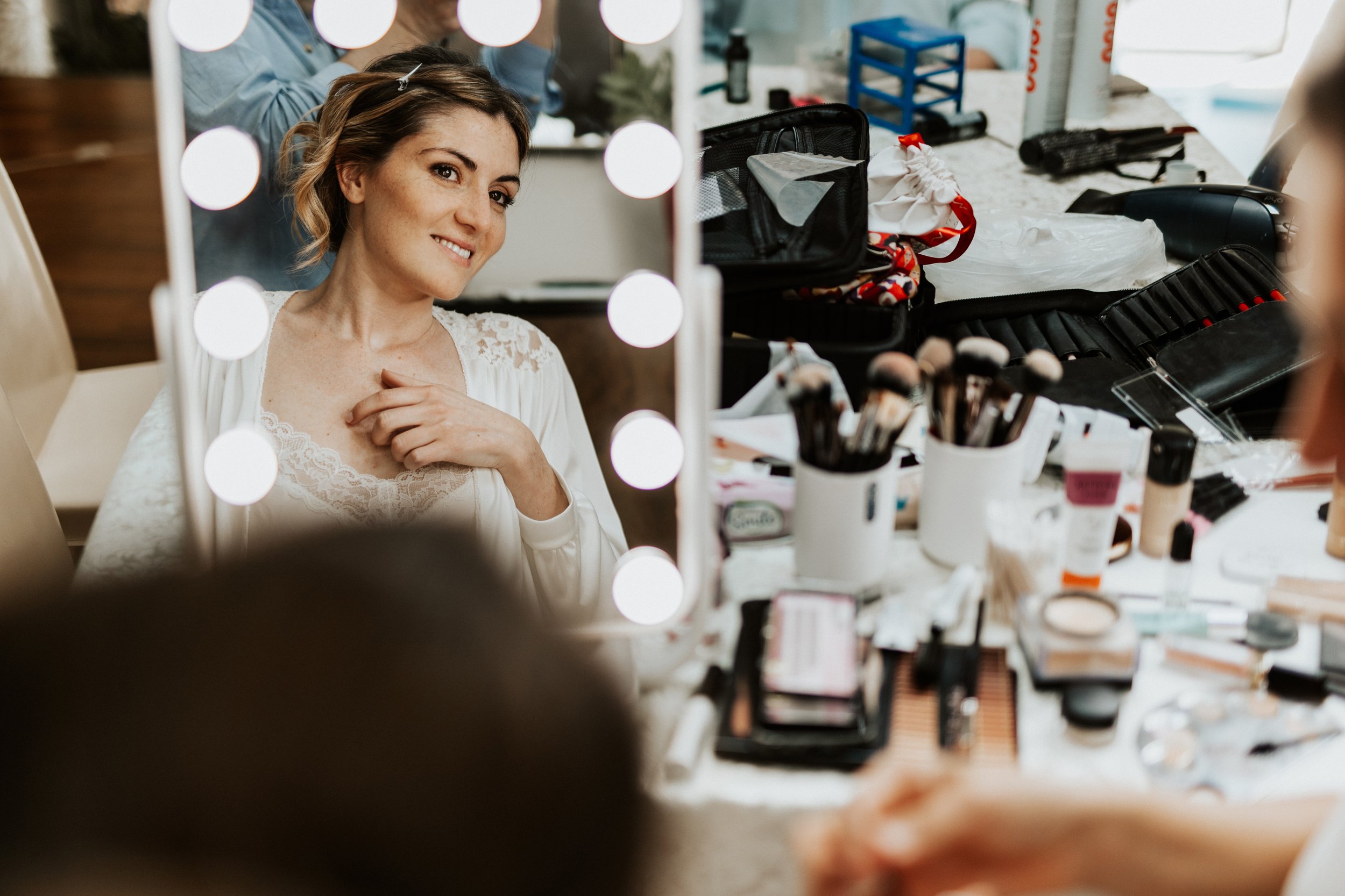 Una donna si guarda allo specchio mentre si prepara, con luci di trucco intorno. Sul tavolo ci sono vari strumenti di trucco e cosmetici.