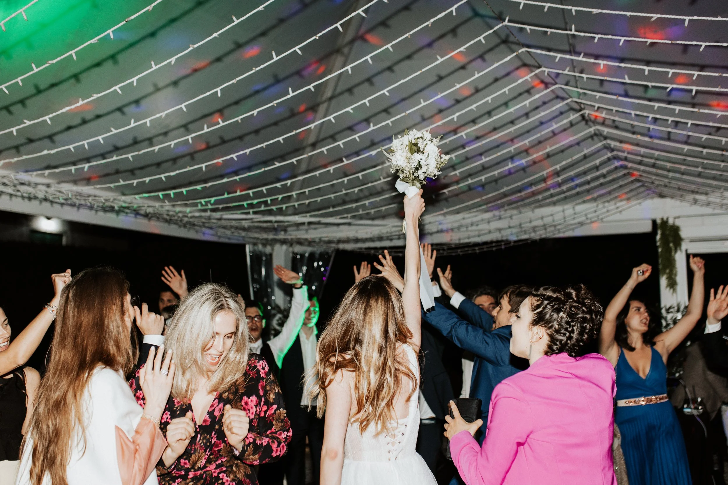 Una festa di matrimonio con persone felici che ballano e una donna con un vestito bianco tiene in alto un bouquet di fiori.