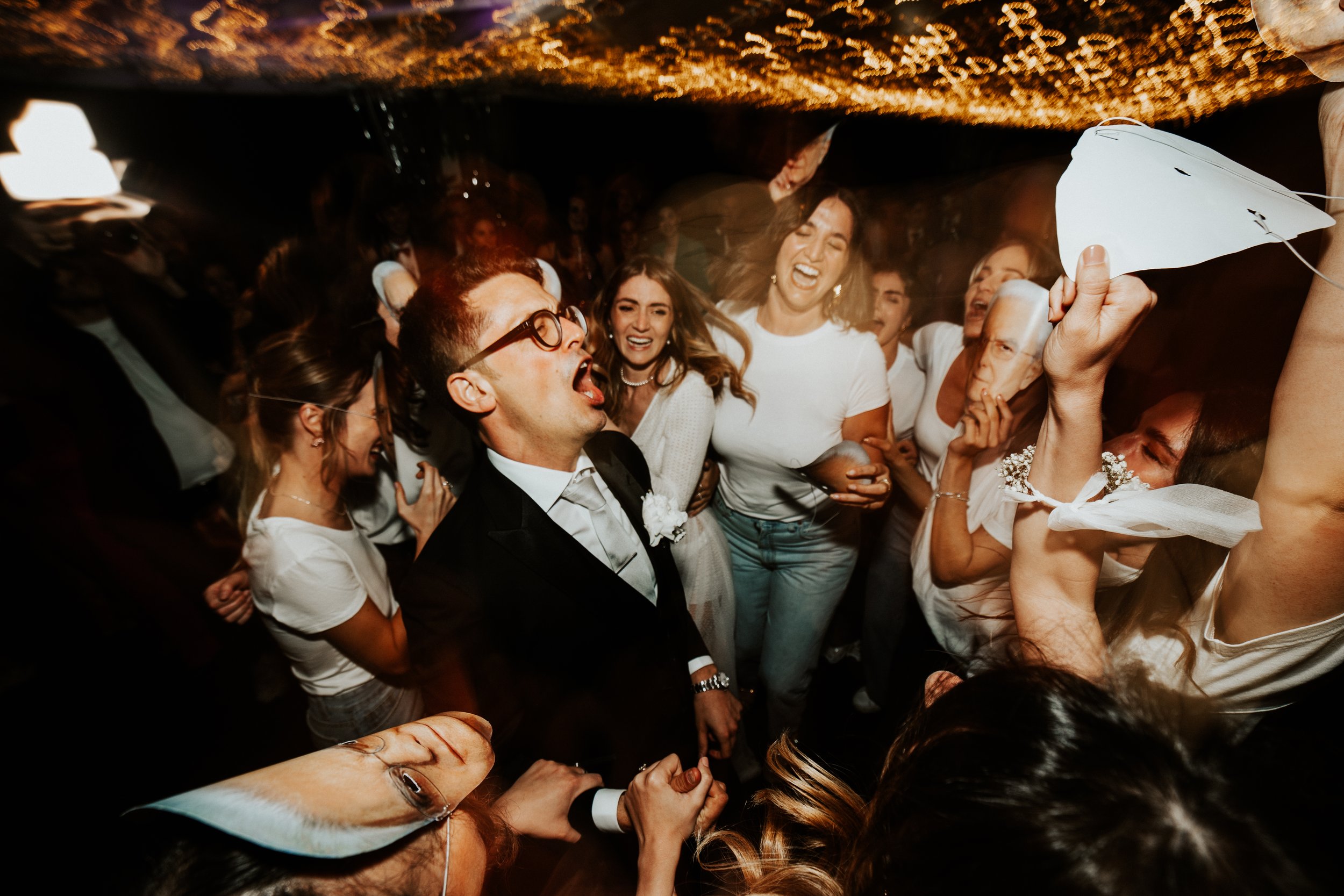 Gruppo di persone felici che ballano e si divertono a una festa, con alcuni che indossano abiti eleganti e altri in abbigliamento casual, sotto luci calde.