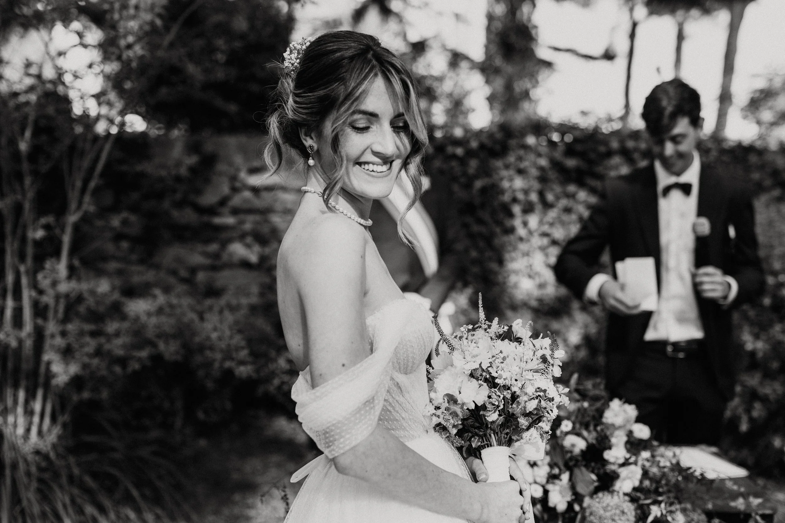 Una donna sorridente in abito da sposa che tiene un bouquet di fiori, durante un matrimonio all'aperto con due uomini vestiti eleganti sullo sfondo.
