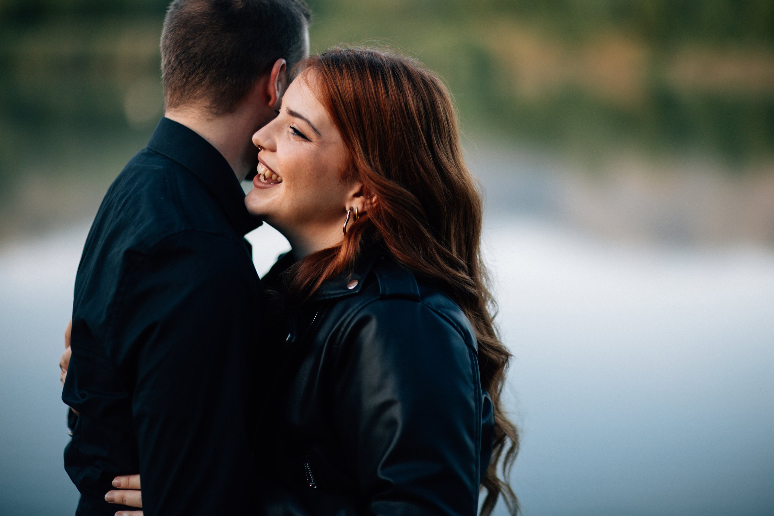 Una coppia che si abbraccia e sorride vicino a un corpo d'acqua, con uno sfondo naturale sfocato.
