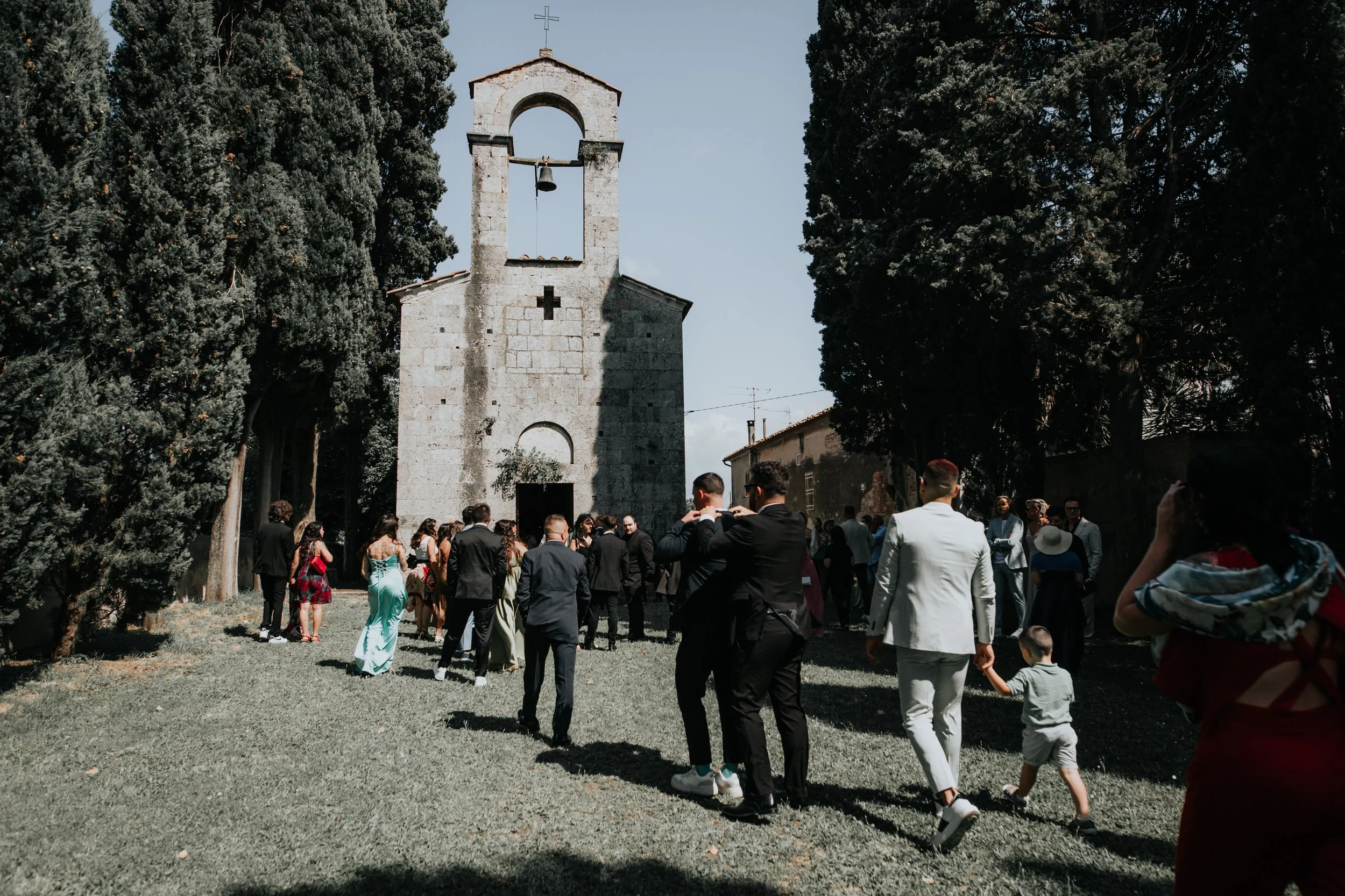 Gruppo di persone vestite elegantemente che si diriggono verso una chiesa in pietra, circondata da alberi, in una giornata soleggiata.