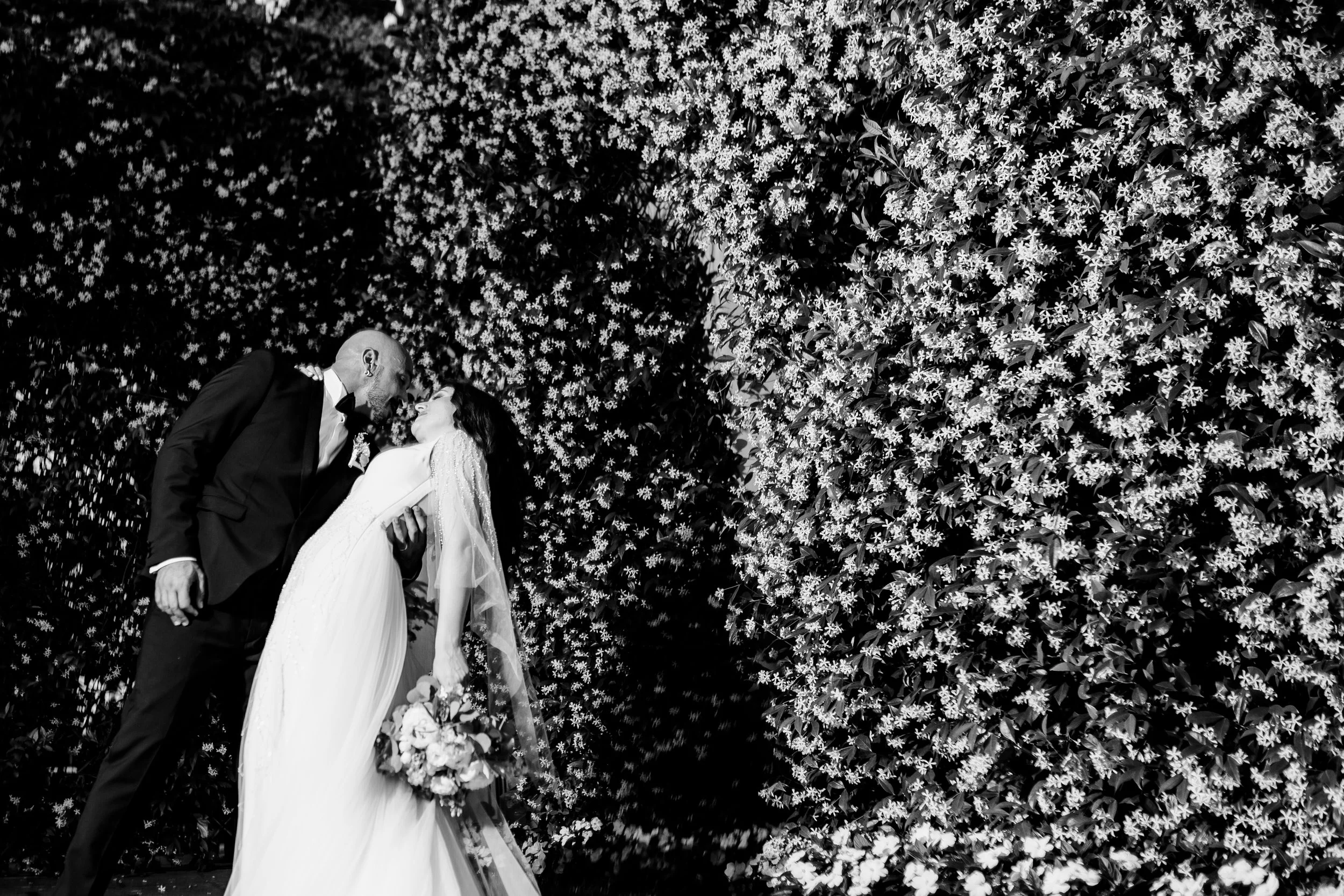 Una coppia di sposi in abiti da matrimonio si bacia davanti a un muro di fiori bianchi.