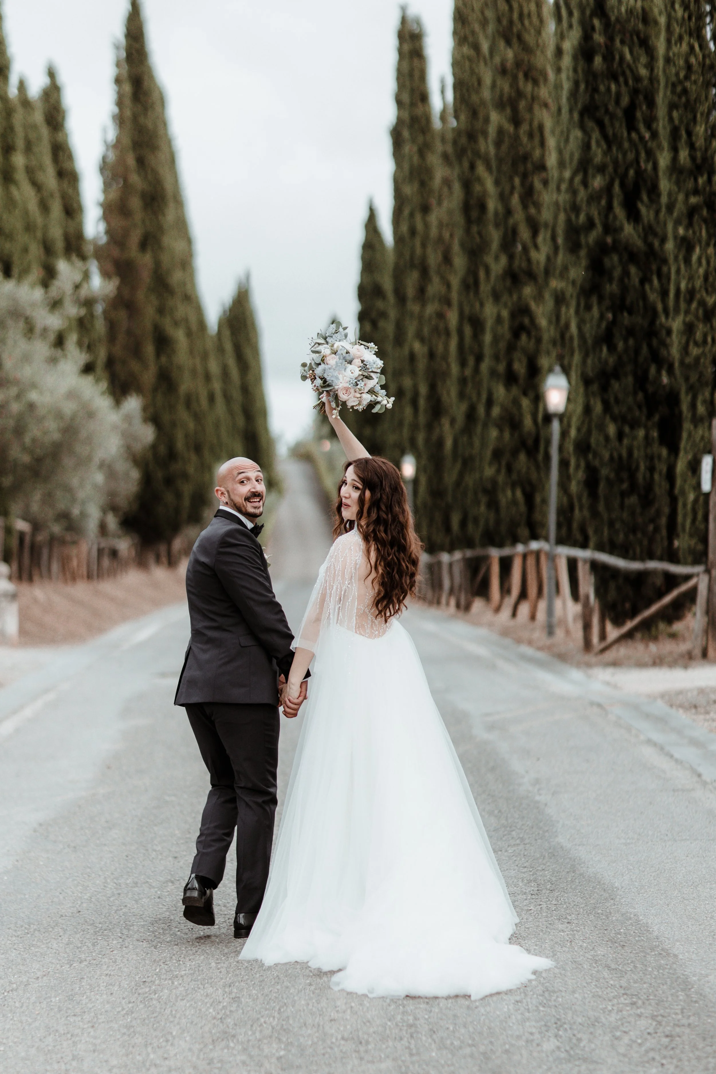 Sposo e sposa sorridenti tra gli alberi lungo una strada campestre, la sposa tiene un bouquet di fiori alzato.