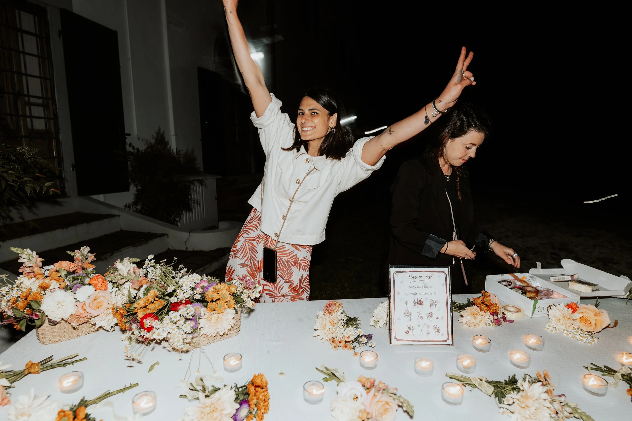 Donna sorridente con braccia alzate, dieta il di fuori, circondata da fiori e candele su un tavolo, in una serata all'aperto.