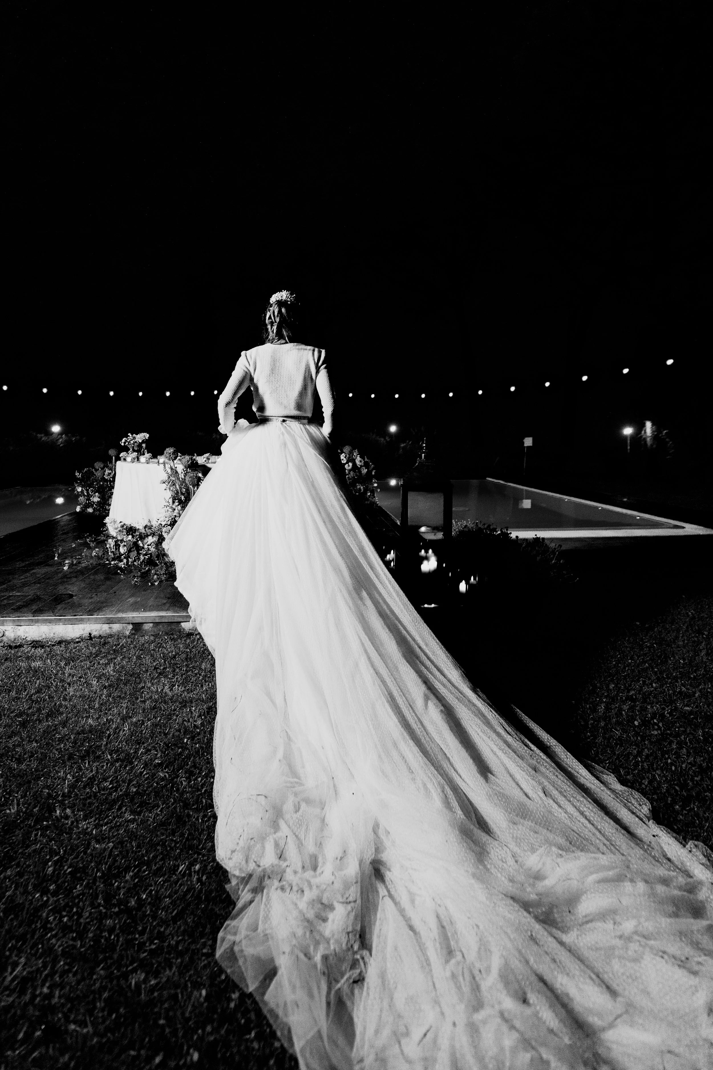 Una sposa in vestito da matrimonio vista da dietro, di notte, con la gonna lunga e il treno che si estende dietro di lei, vicino a una piscina e un tavolo con fiori.