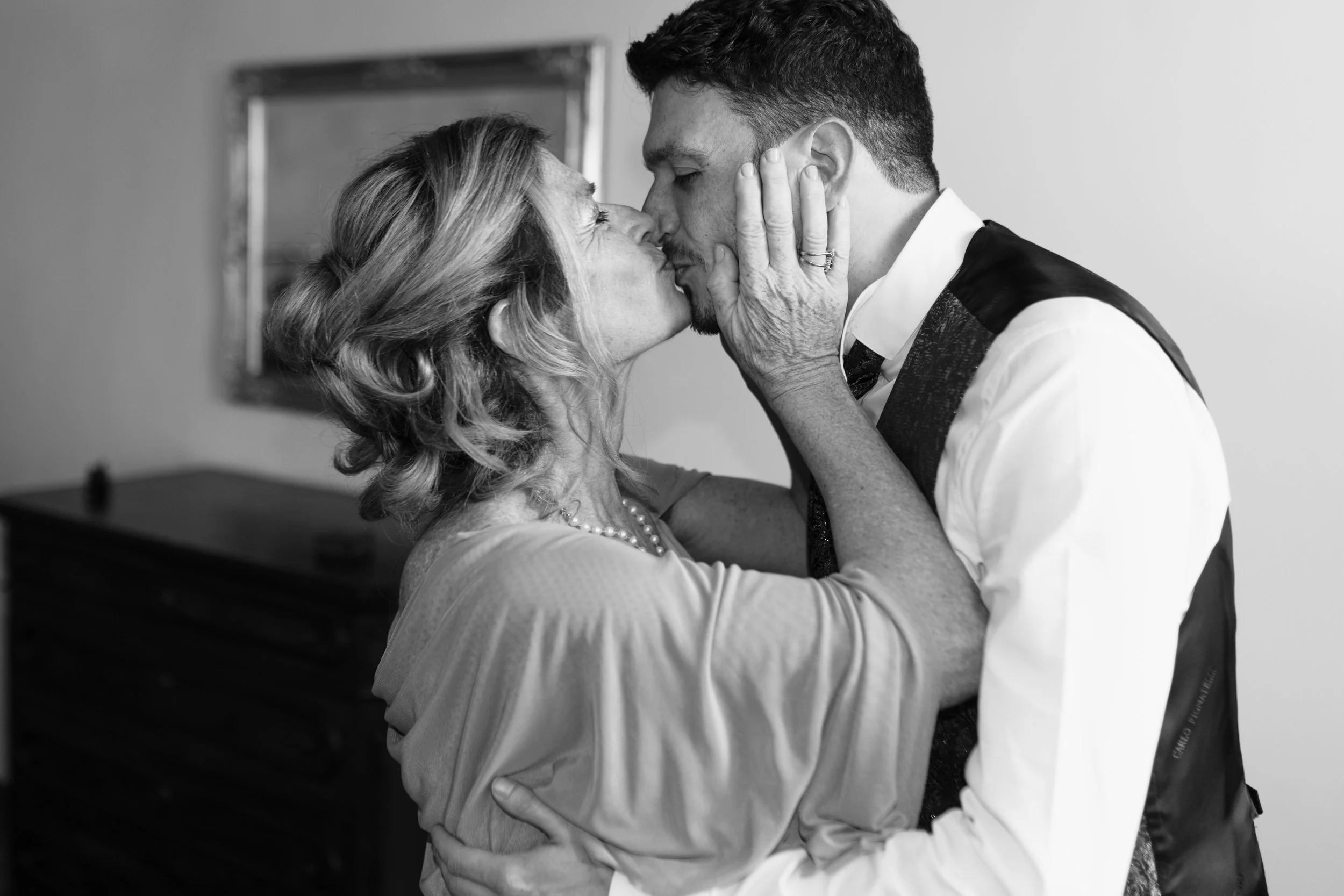 Una donna anziana e un uomo giovane si scambiano un bacio affettuoso, toccandosi il volto, in una stanza