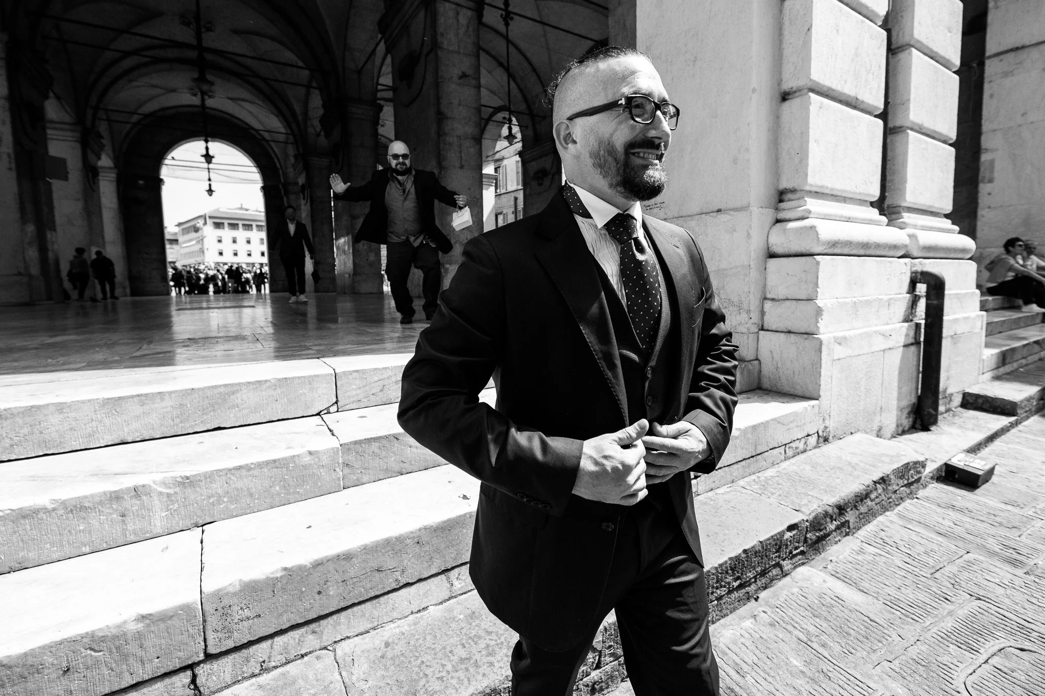 Uomo in abito elegante con papillon, occhiali e barba, sorride mentre cammina davanti a un edificio storico, altri uomini in background