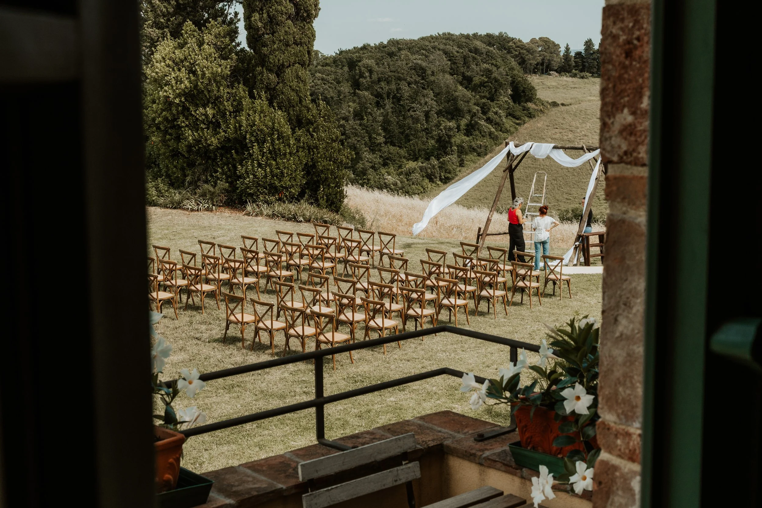 Scena all'aperto di preparativi per un matrimonio con sedie attorno a un'area decorata, vista attraverso una finestra con fiori bianchi. Due persone si trovano vicino a una struttura drappeggiata in tessuto bianco. Sullo sfondo si vedono verdi collin
