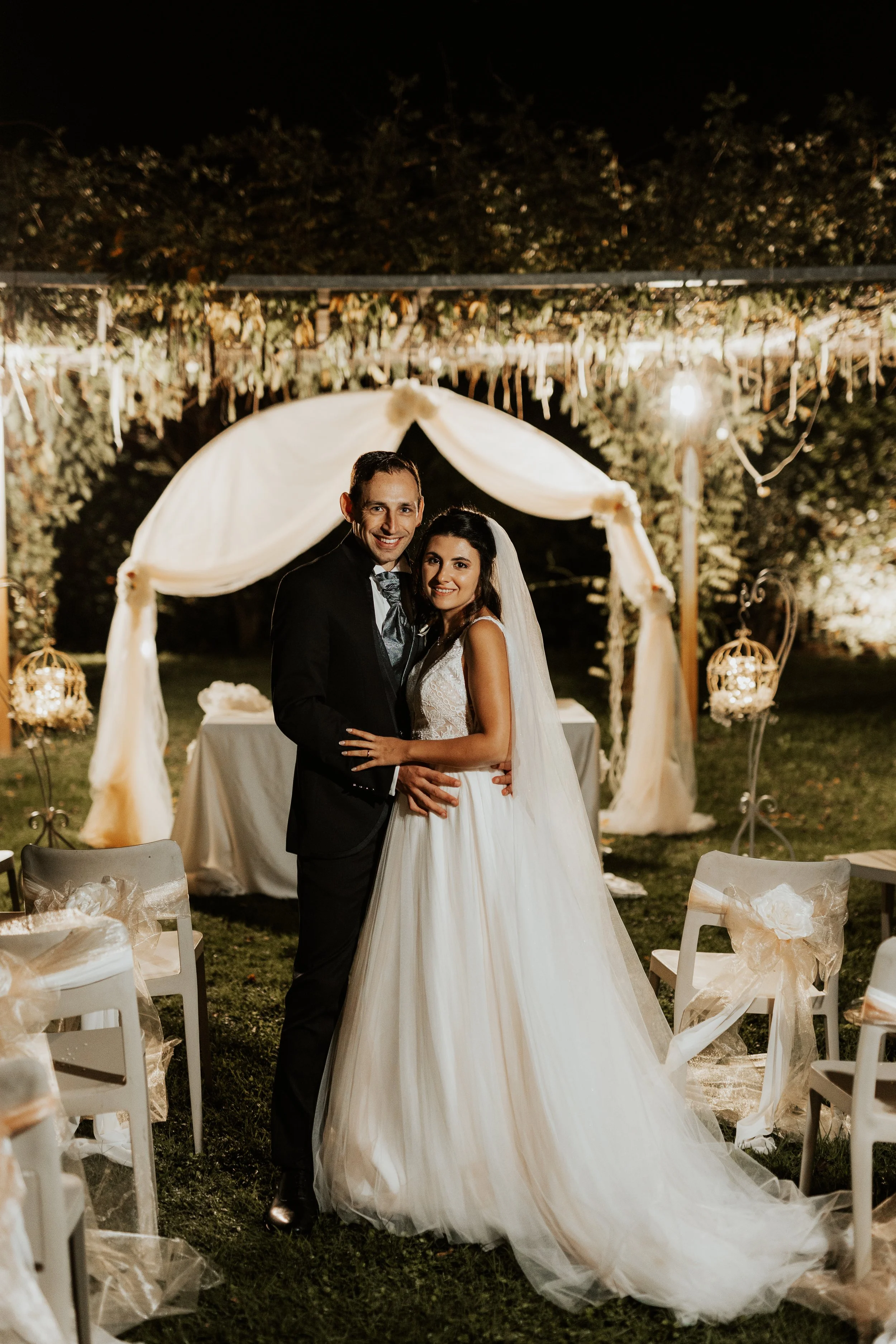 Foto di una sposa e uno sposo che sorridono, abbracciati, durante il loro matrimonio all'aperto di sera, sotto una luce calda e decorazioni floreali.