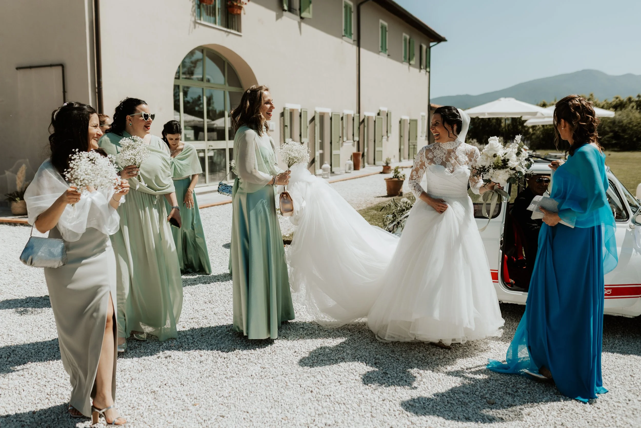 Gruppo di donne in abiti eleganti e una sposa in abito bianco con un bouquet che ride e socializza all'aperto, vicino a una macchina d'epoca, con un edificio e un paesaggio montano sullo sfondo.