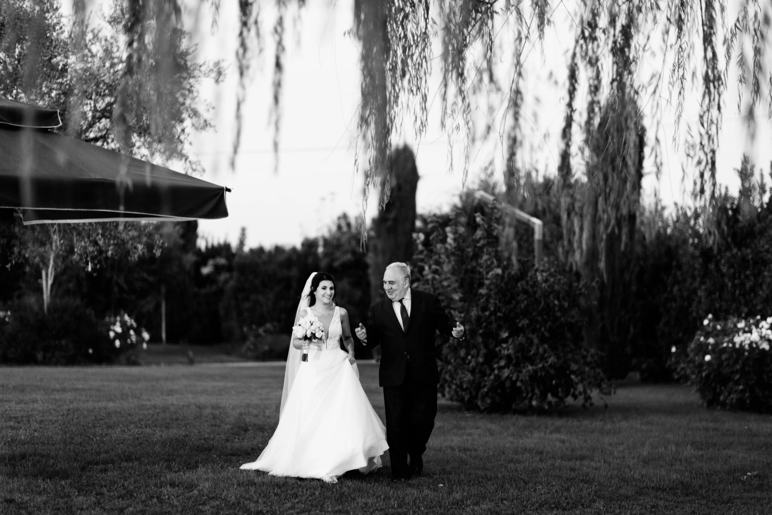 Foto in bianco e nero di una sposa e un uomo, probabilmente il padre, che camminano insieme sorridenti all'aperto in un giardino, in occasione di un matrimonio.