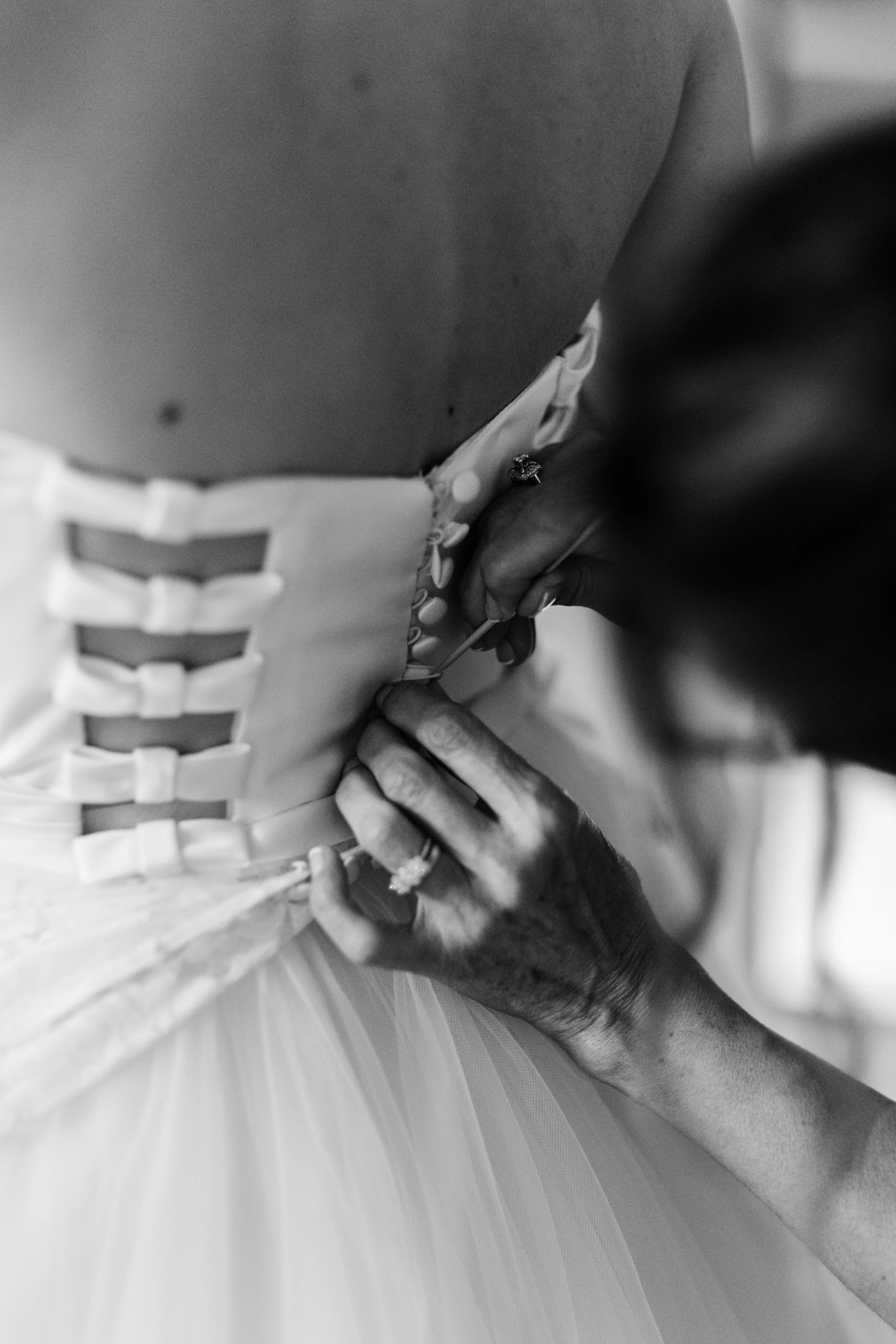 Una persona sta cucendo i bottoni sulla schiena di un abito da sposa bianco, in un'immagine in bianco e nero.