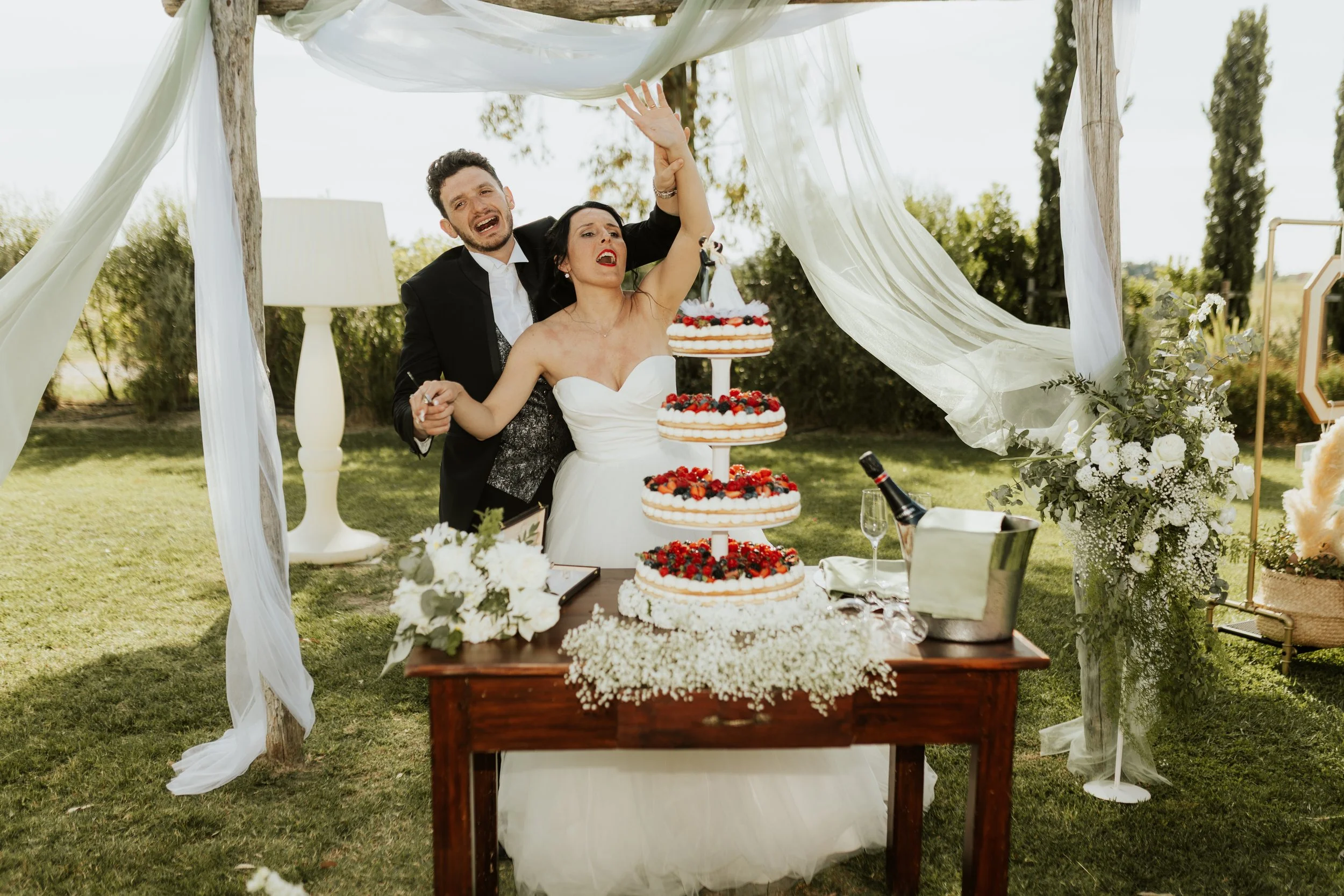 Una coppia di sposi festeggia il matrimonio davanti a una torta nuziale all'aperto, circondati da decorazioni floreali e drappeggi di stoffa bianca.