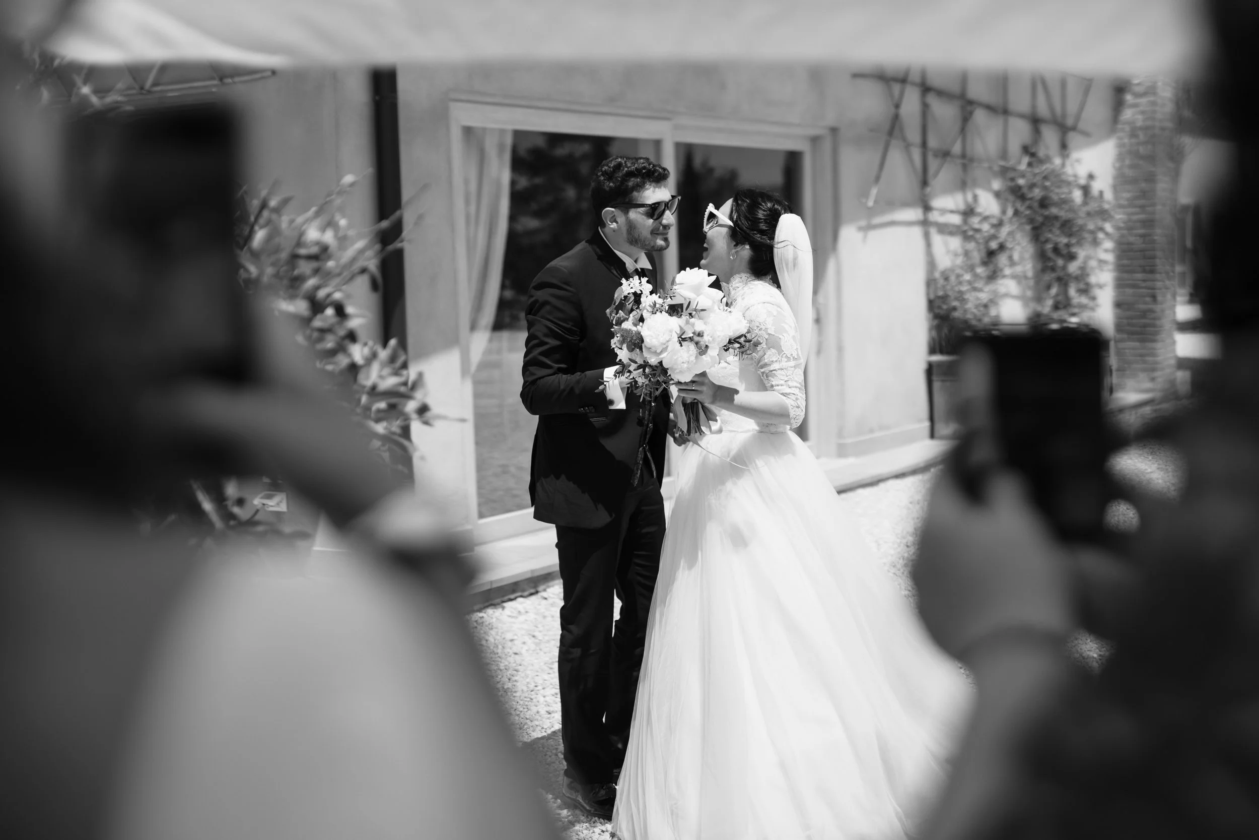 Una coppia di sposi si abbraccia e si sorride davanti a una casa, con fianco il volto di alcune persone che li fotografano.