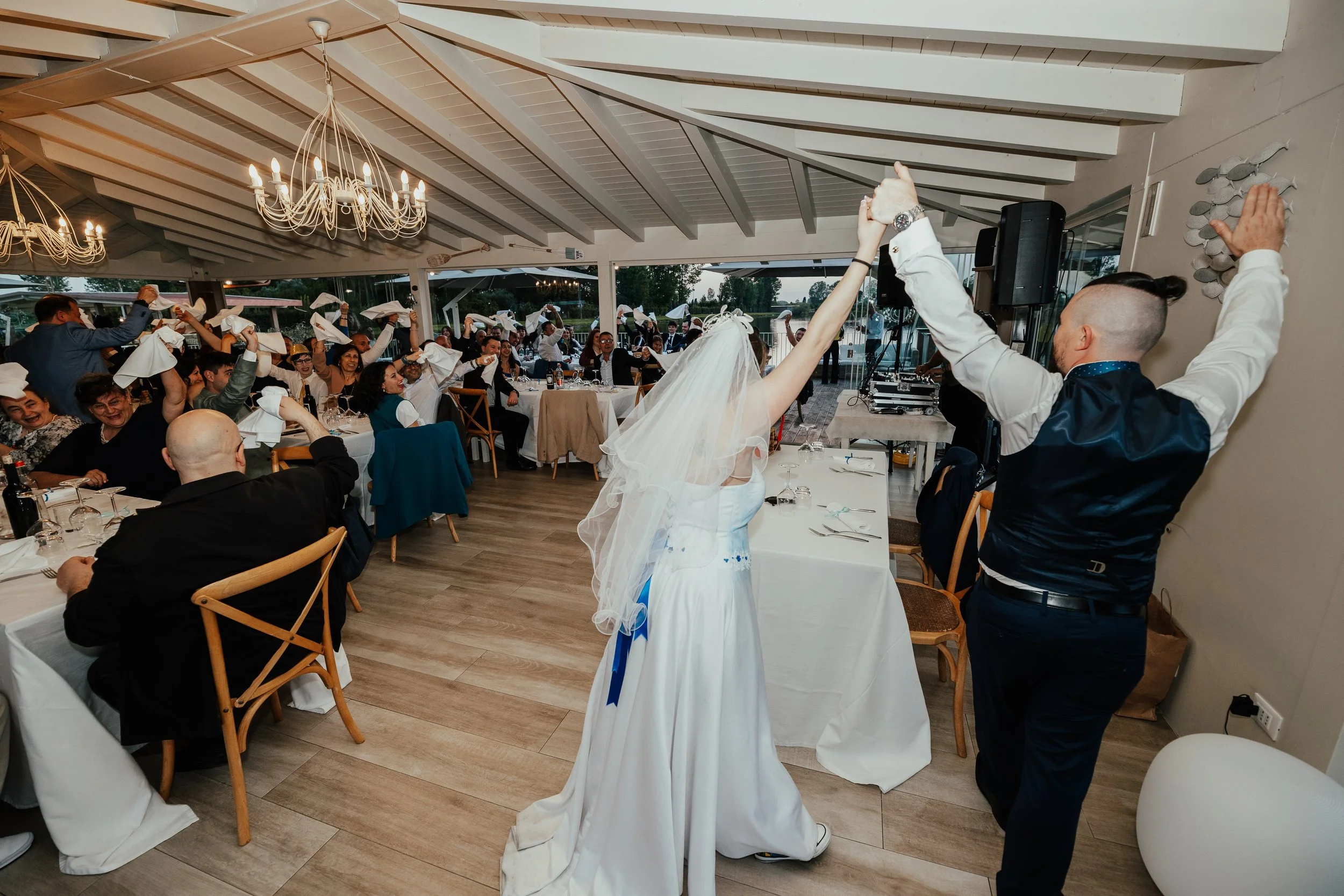 Una coppia di sposi balla con gli invitati seduti a tavola in un ricevimento di nozze in un ristorante con soffitto a travi di legno e lampadari. L'atmosfera è festosa e allegra.