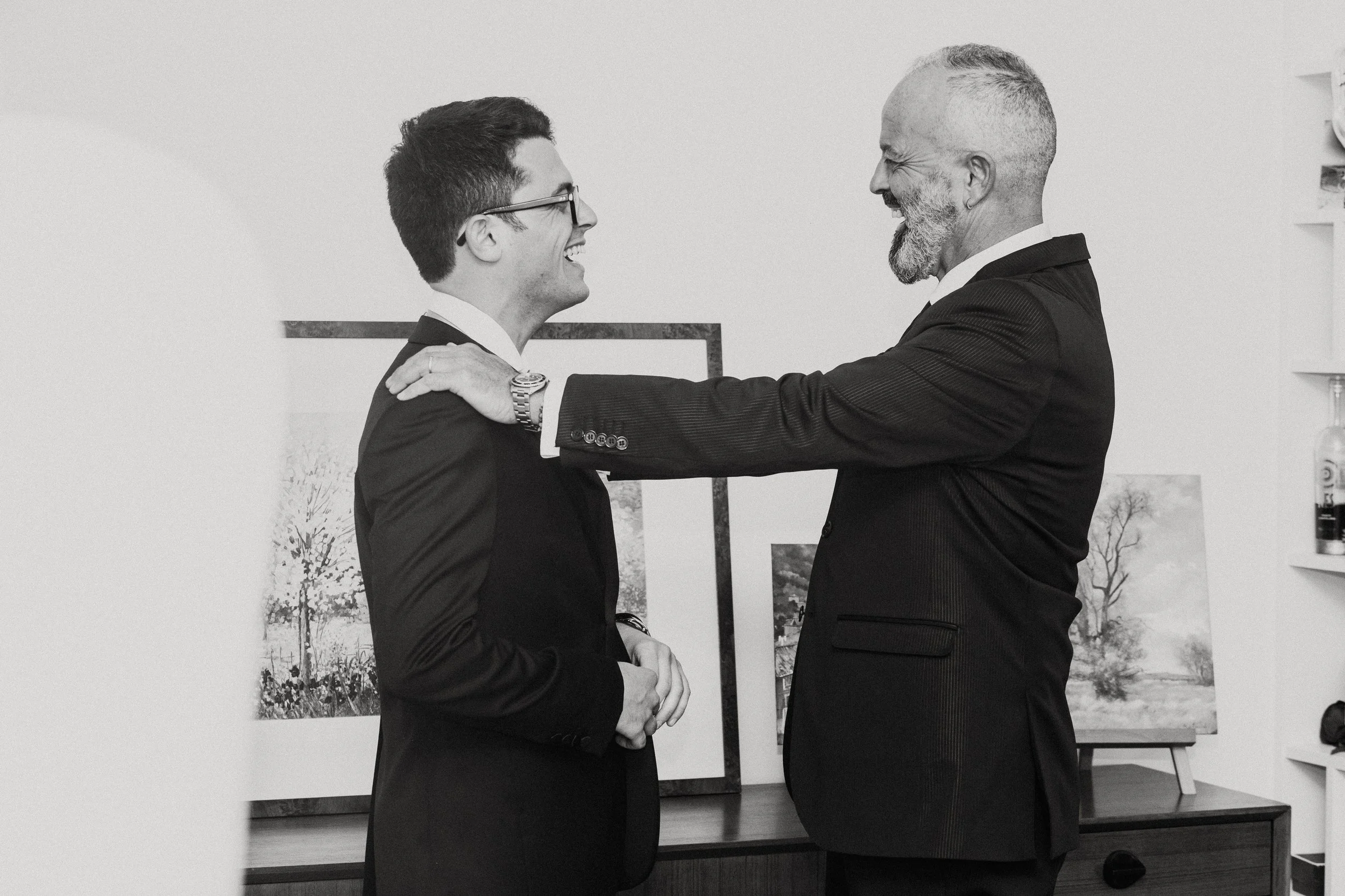 Un uomo giovane con gli occhiali e un uomo anziano sorridenti si abbracciano in un ambiente con quadri alle pareti.