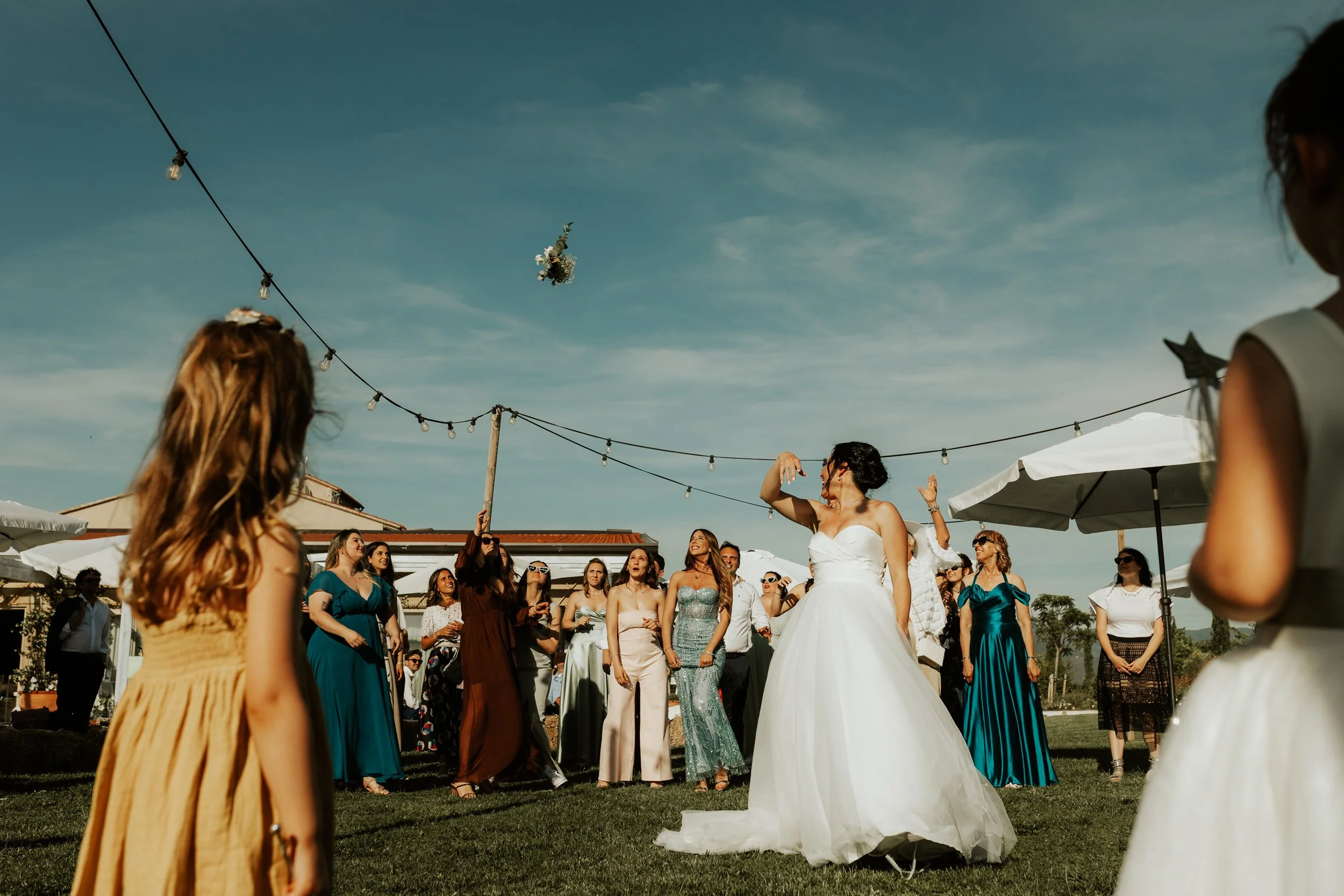 Una coppia di sposi che si abbracciano durante un matrimonio all'aperto, circondata da amici e parenti, con decorazioni di luci e un cielo blu chiaro.