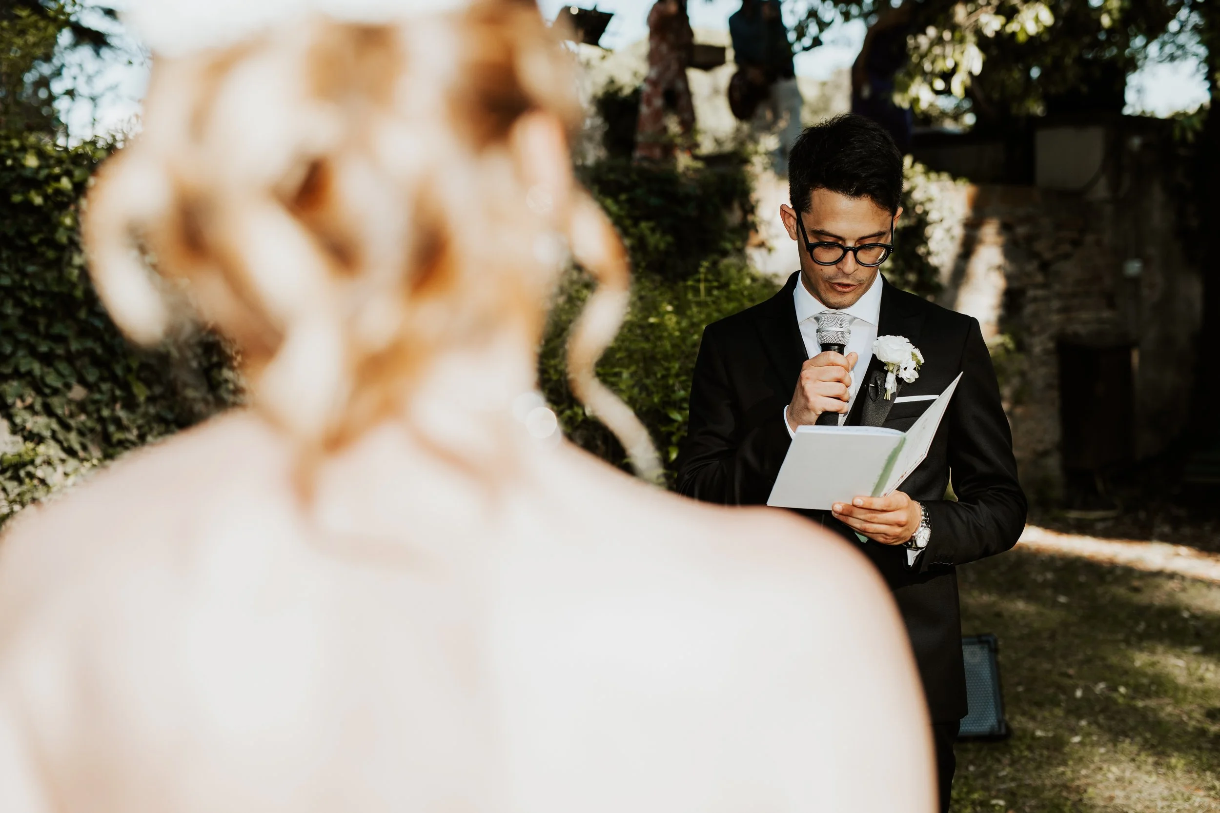 Uomo in abito nero con fiocco bianco e occhiali, che legge da un foglio durante un matrimonio all'aperto, con una sposa sfocata in primo piano.