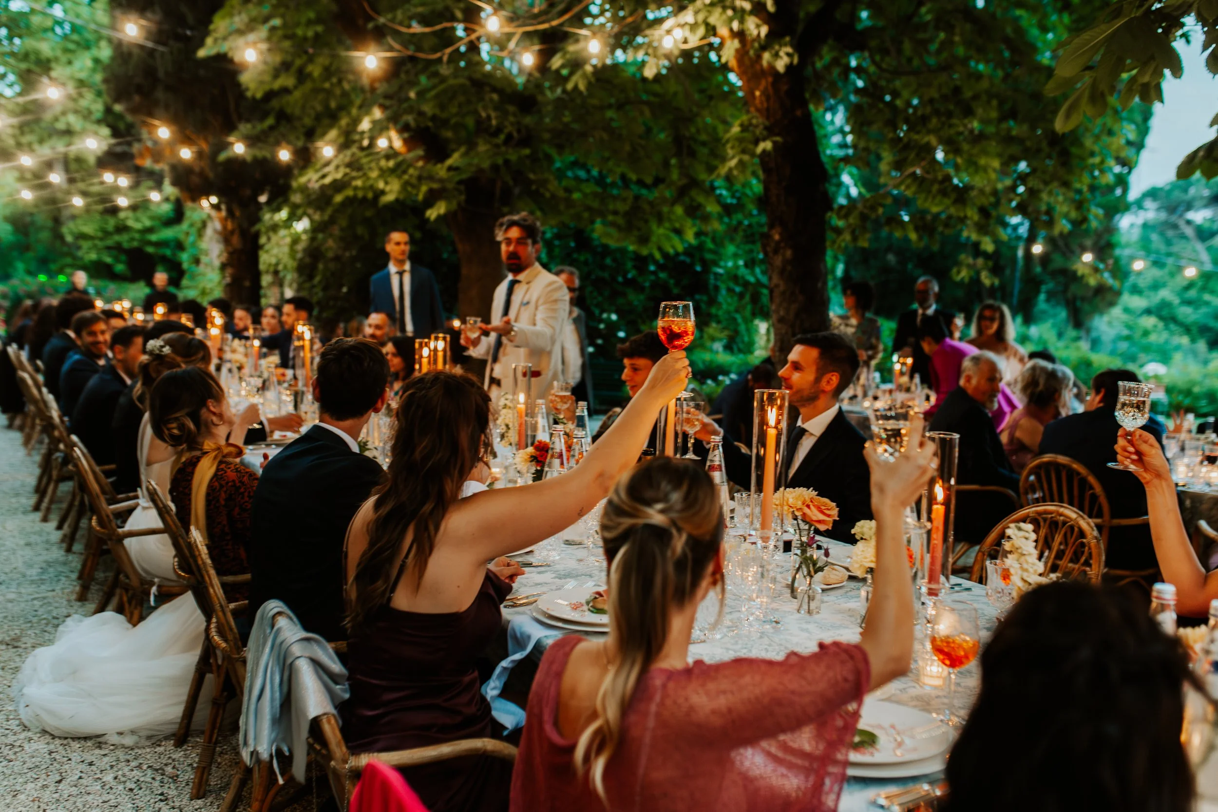 Ricevimento in giardino con persone sedute intorno a un lungo tavolo decorato con candele e fiori, tra alberi e luci sospese, persone che brindano dall'atmosfera festosa.