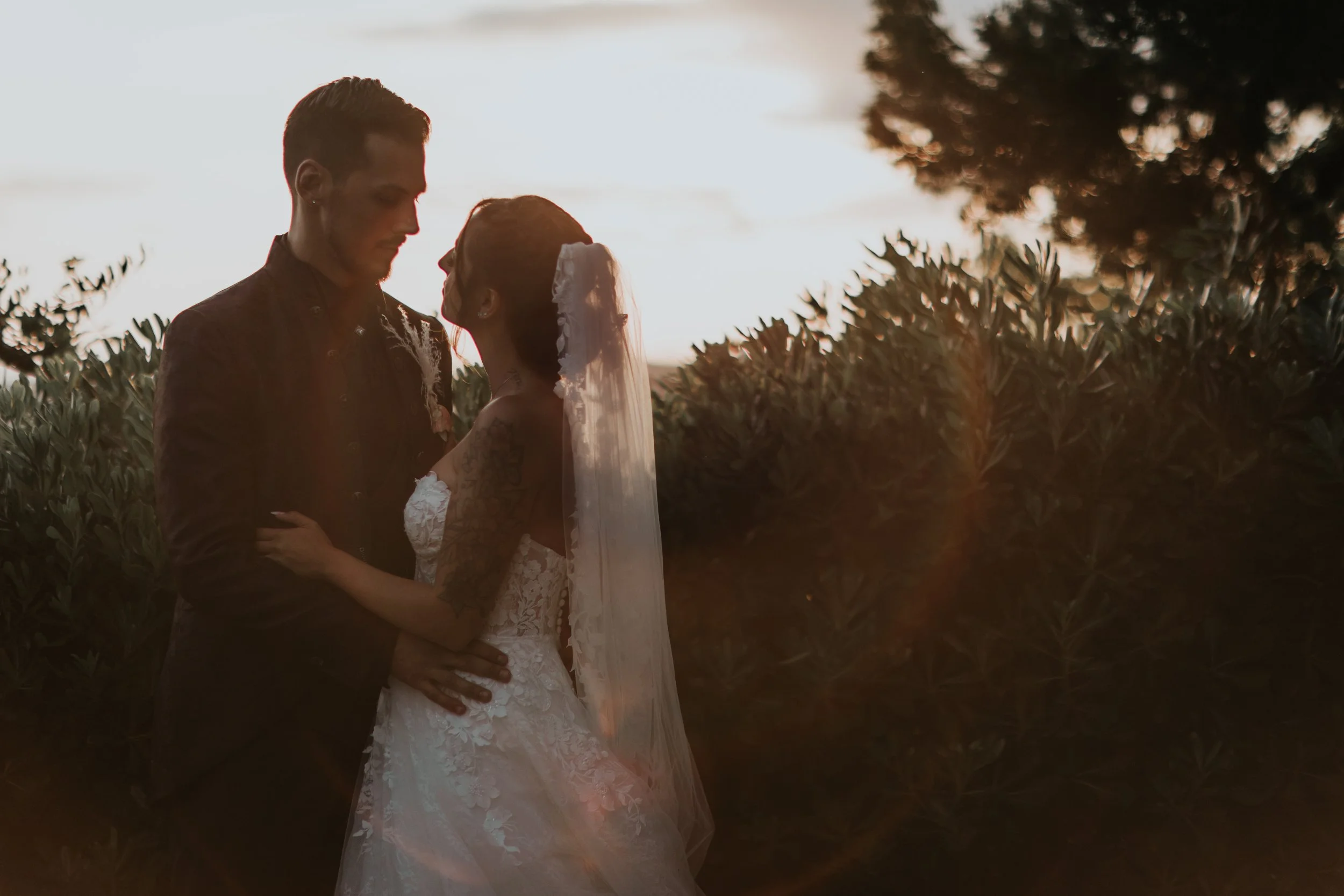 Una coppia di sposi si guarda intimi all'aperto durante il tramonto, circondati da vegetazione.
