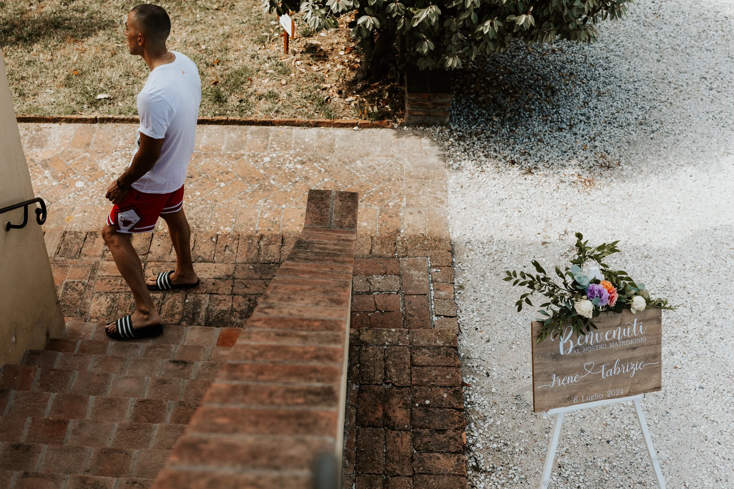 Uomo vestito in tenuta casual con maglietta bianca, pantaloncini rossi e ciabatte adatte alla spiaggia, sale le scale di pietra in un ambiente esterno. Accanto, un cartellone di benvenuto con fiori colorati e scritte in italiano, che dice 'Benvenuti